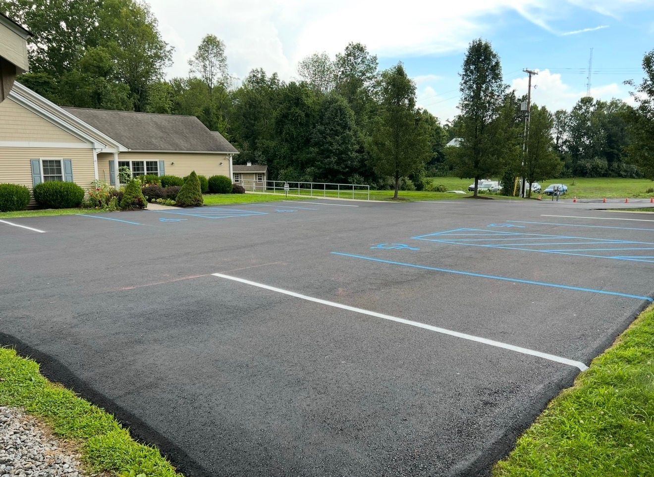 A newly paved asphalt parking lot with white and light blue parking space markings, next to a building and greenery.