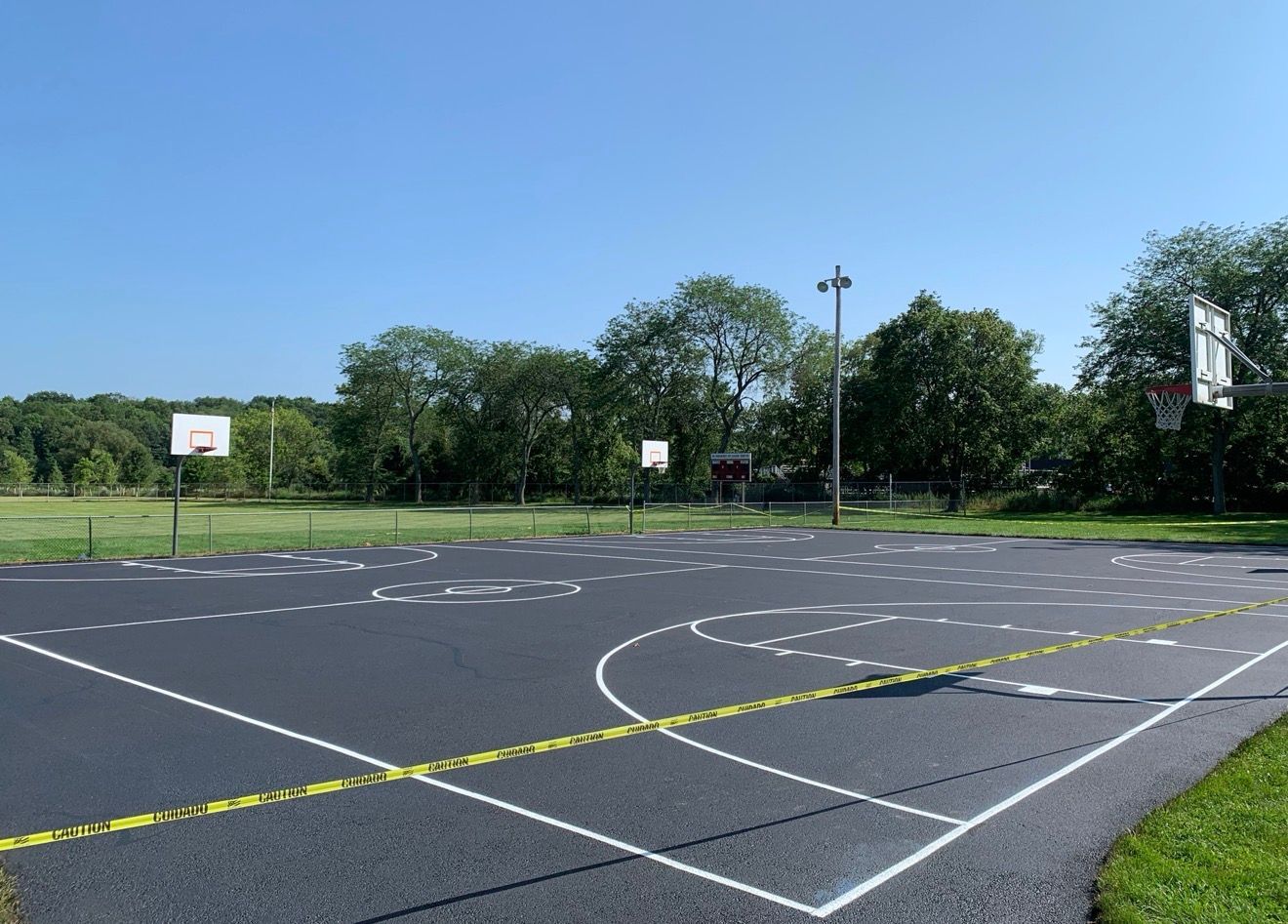 A freshly paved outdoor basketball court with white markings on a clear day, surrounded by green trees and grass.