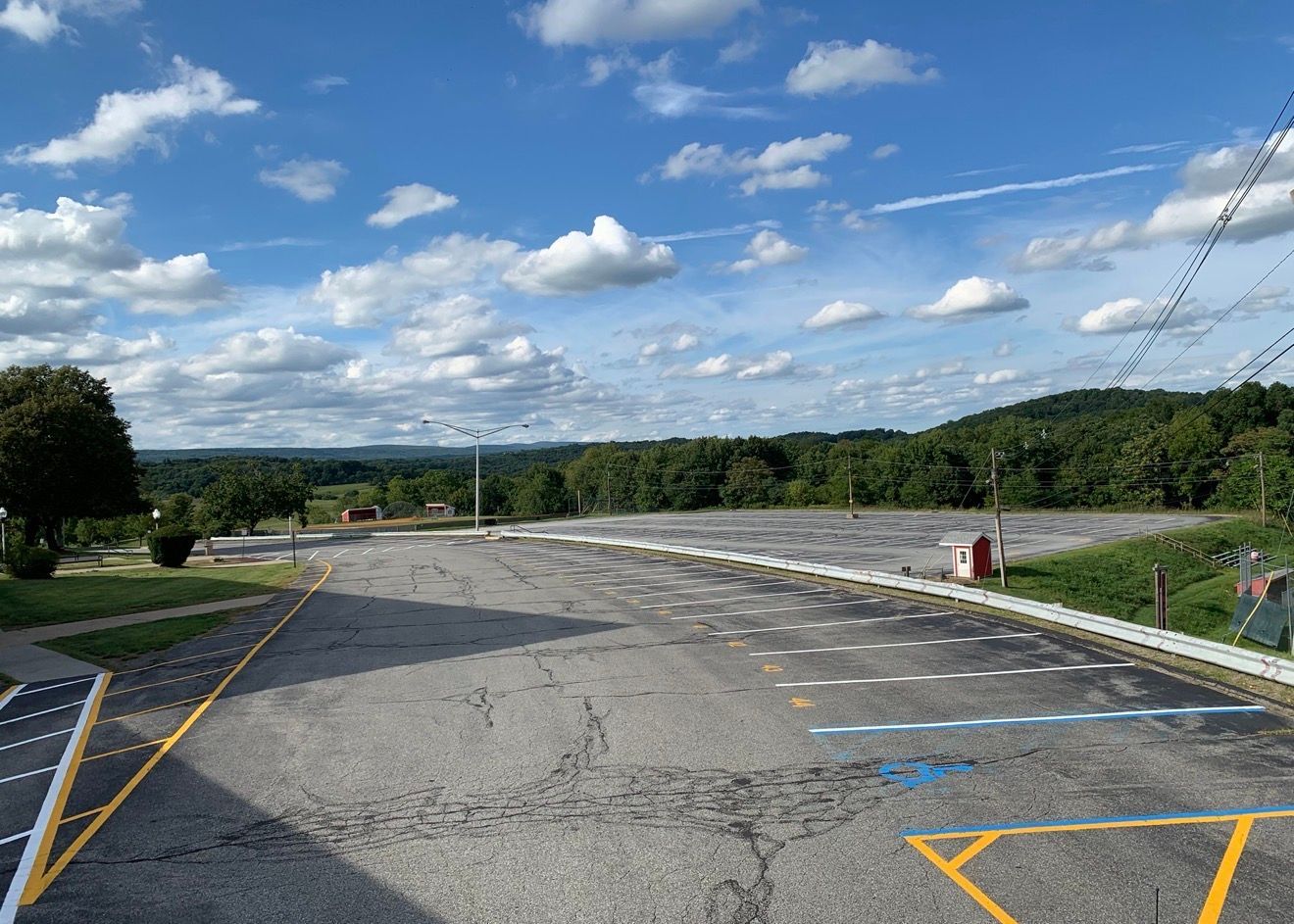 An empty paved parking lot with painted stalls under a blue sky with white clouds, bordered by trees on the horizon.