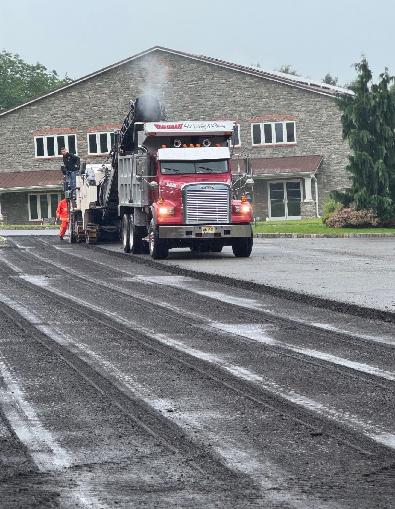 A red dump truck and road milling machine operate on a newly milled asphalt parking lot in front of a stone building.
