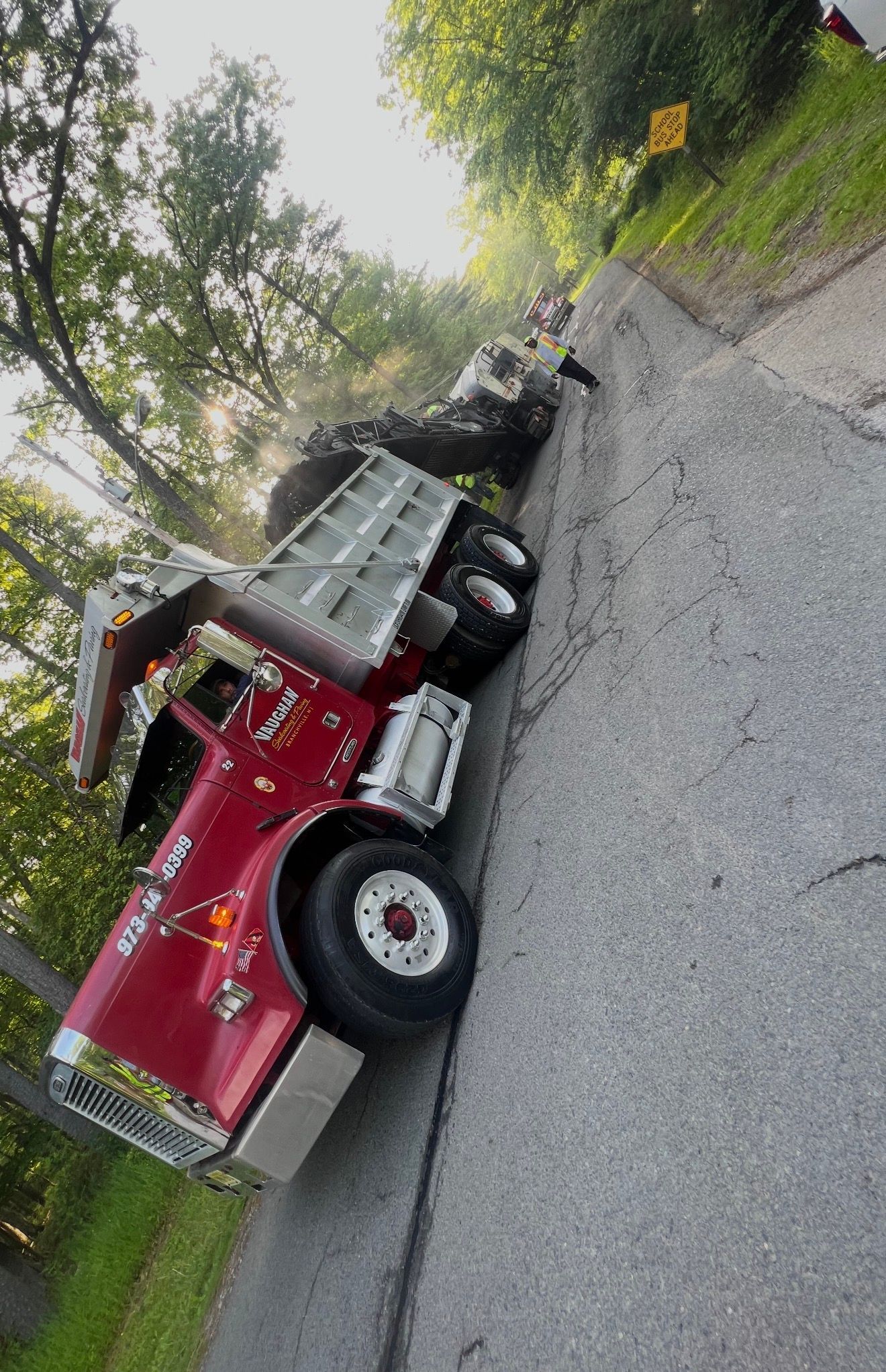 A red dump truck parked on a narrow, asphalt road lined with trees and a yellow sign in the background.