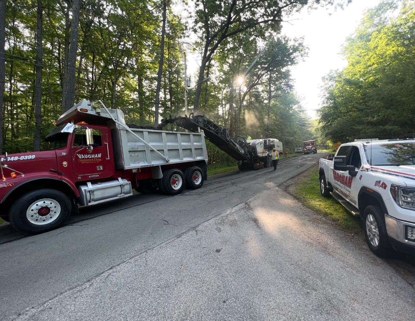 A red dump truck and a construction machine perform road repairs on a tree-lined street with a white utility truck nearby.