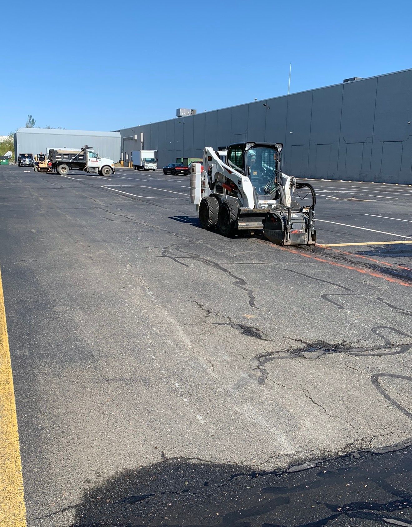 A white skid steer with a concrete saw attachment works on a large asphalt parking lot near a commercial building.