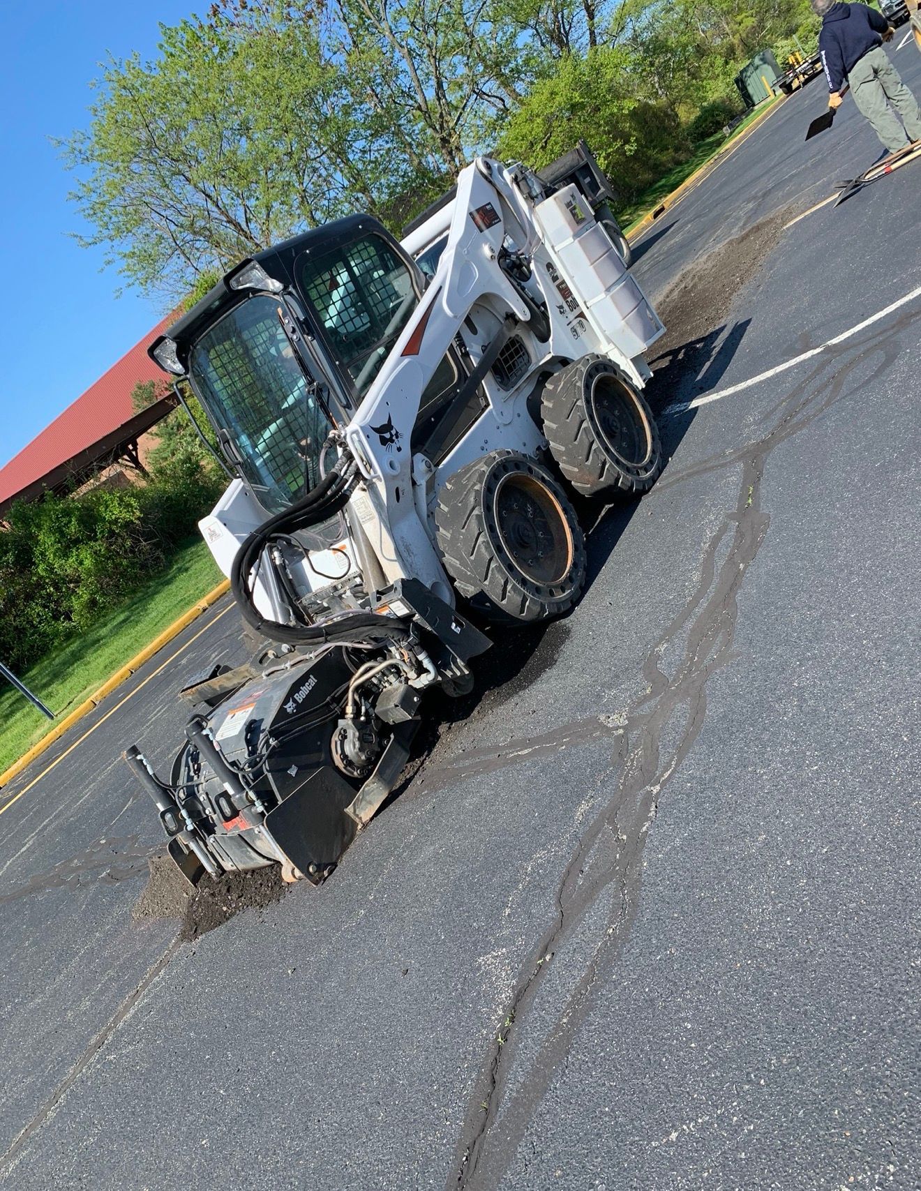 A white Bobcat skid-steer loader with a pavement milling attachment is actively cutting into an asphalt parking lot.