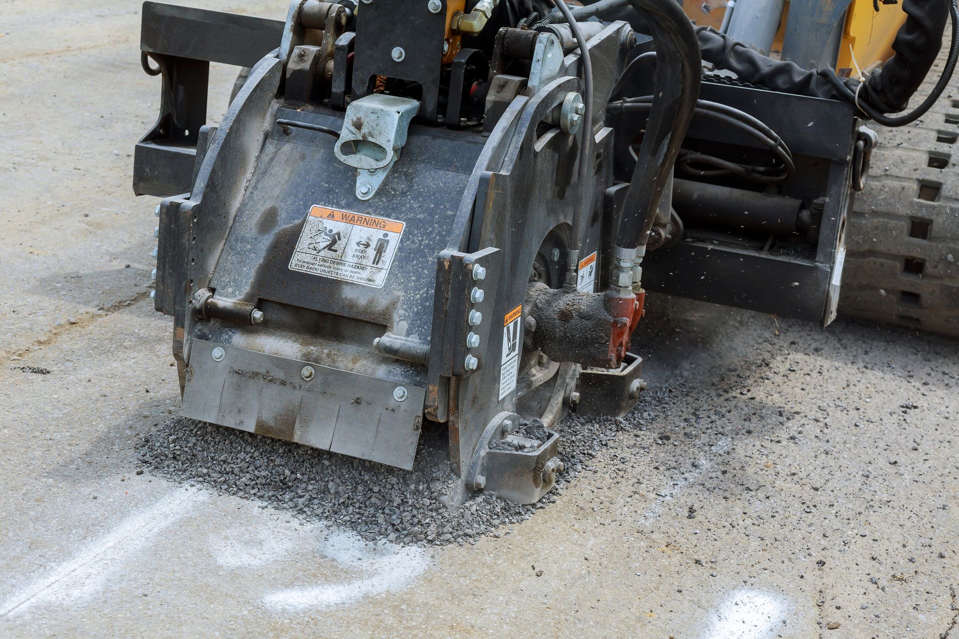 A heavy-duty milling machine attachment mounted on a vehicle cuts into a paved asphalt surface.