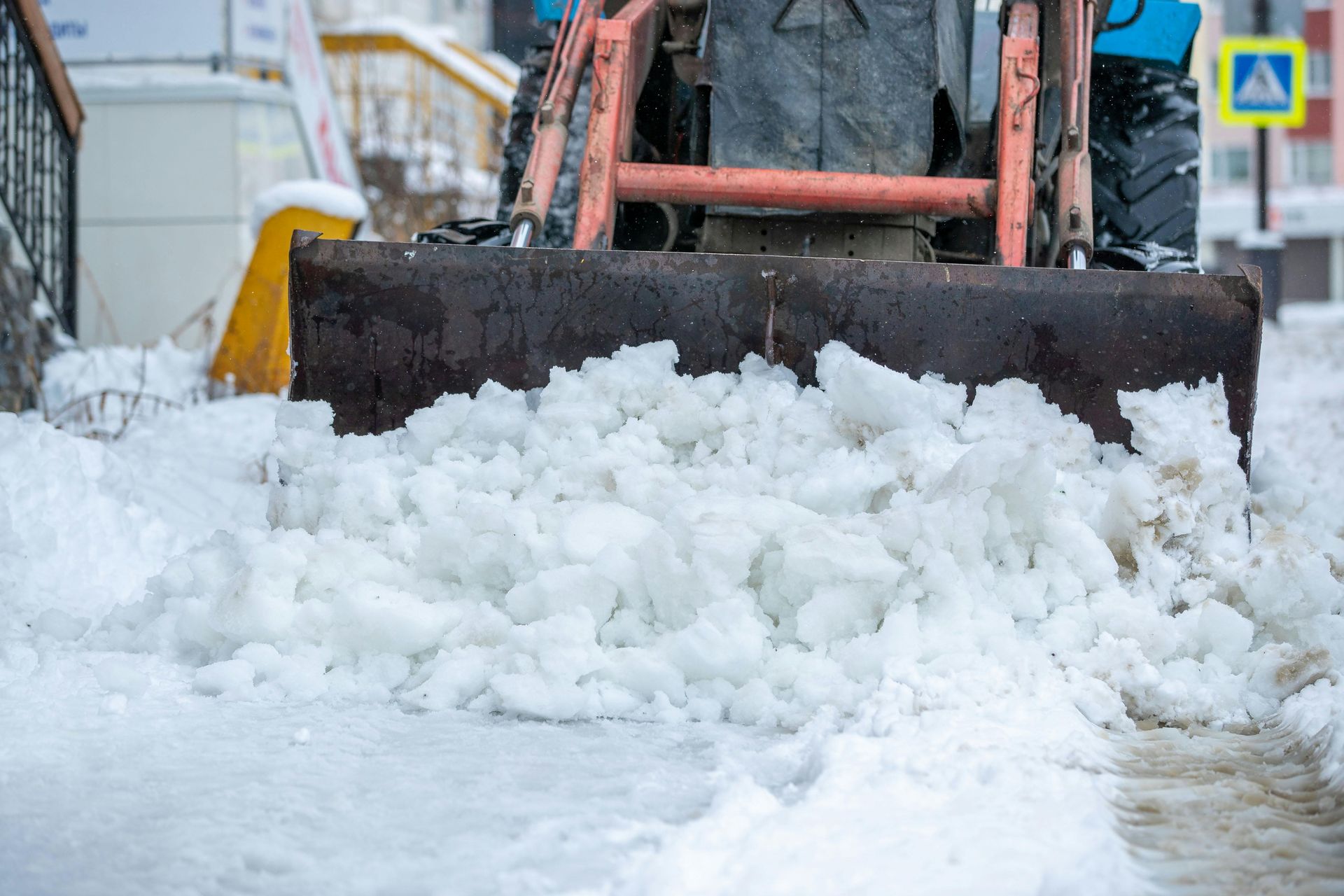 A tractor with a large metal blade pushes a pile of snow and ice across a snowy urban street.