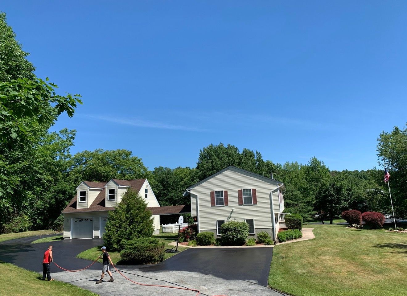 Two people apply sealcoating to a residential driveway in front of a light-colored two-story house on a sunny day.