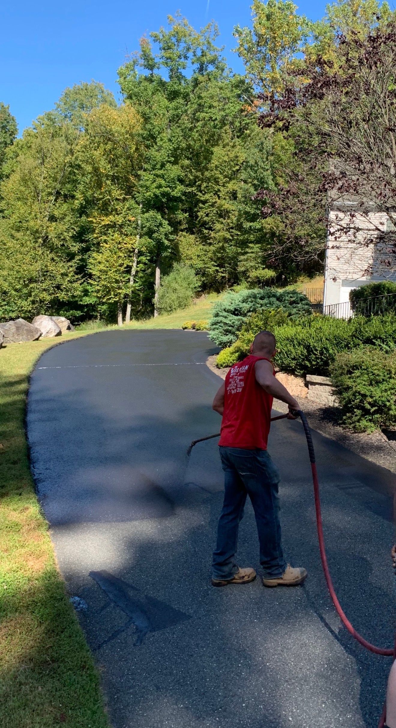 A worker in a red shirt uses a spray nozzle to apply sealant to a residential asphalt driveway.