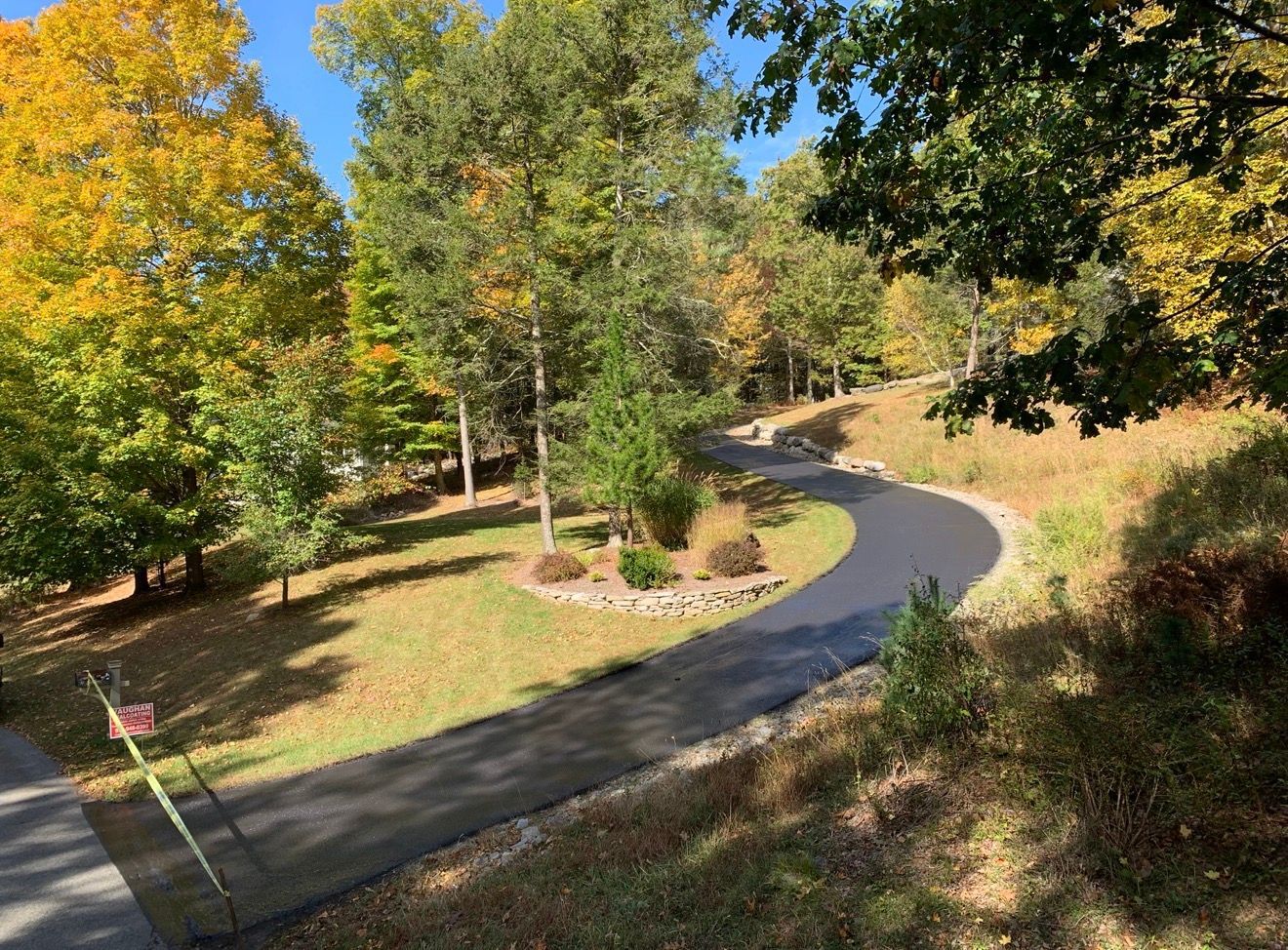 A curved, freshly paved asphalt driveway winds through a wooded residential yard with autumn foliage in shades of yellow.