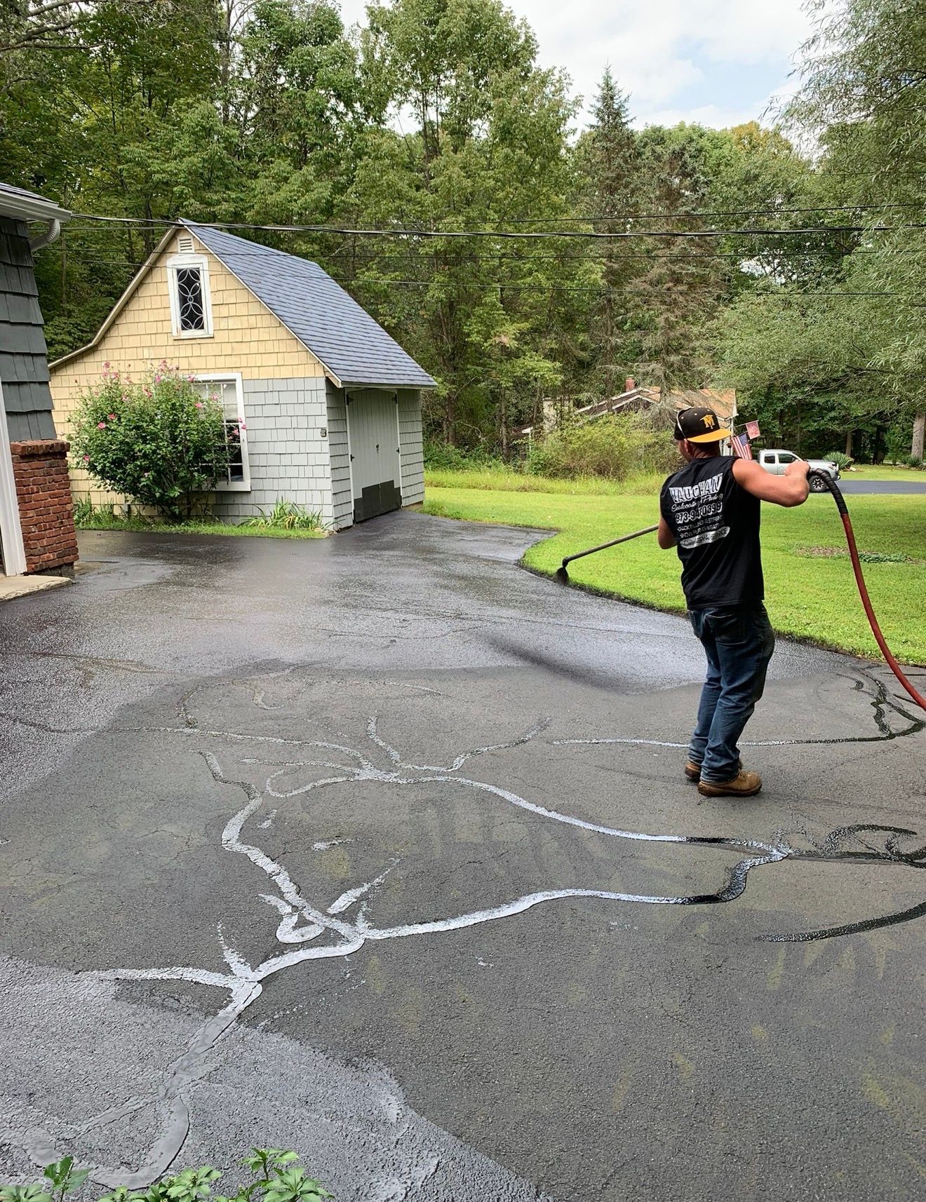 A person in a black shirt uses a pressure washer to clean a dark asphalt driveway in front of a small garage.