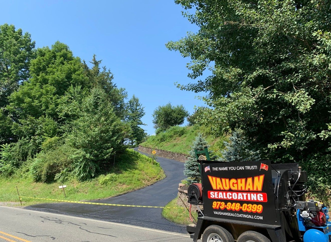 A Vaughan Sealcoating trailer parked next to a newly paved, curving driveway surrounded by trees on a sunny day.