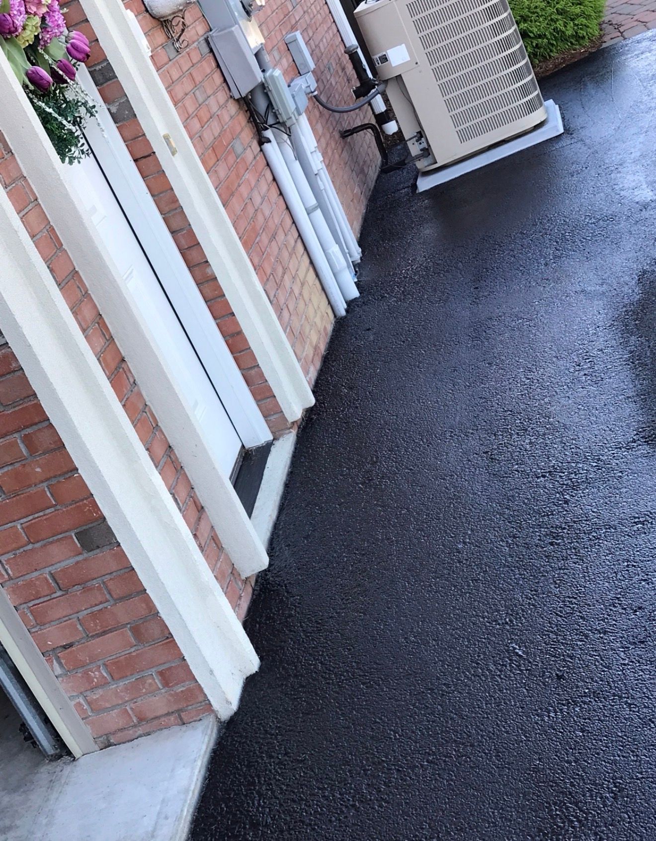 A brick wall with white trim and electrical conduit next to an outdoor HVAC unit on a wet paved surface.