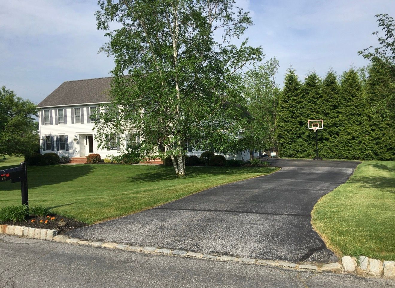 A white two-story house with a long, paved driveway and a basketball hoop, surrounded by green lawns and trees.