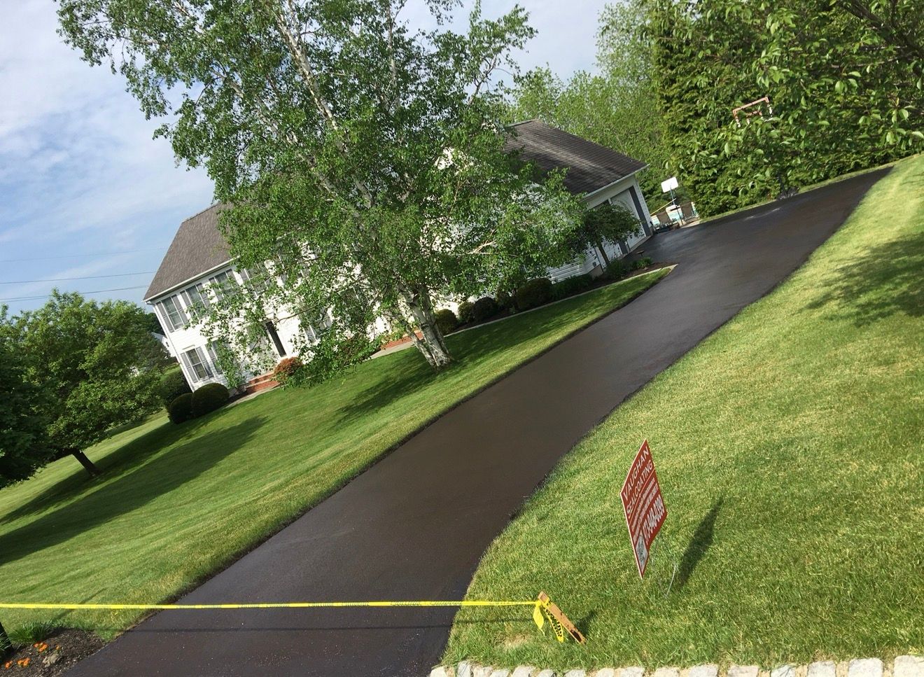 A paved driveway leads to a white house surrounded by a green lawn and trees on a sunny day.