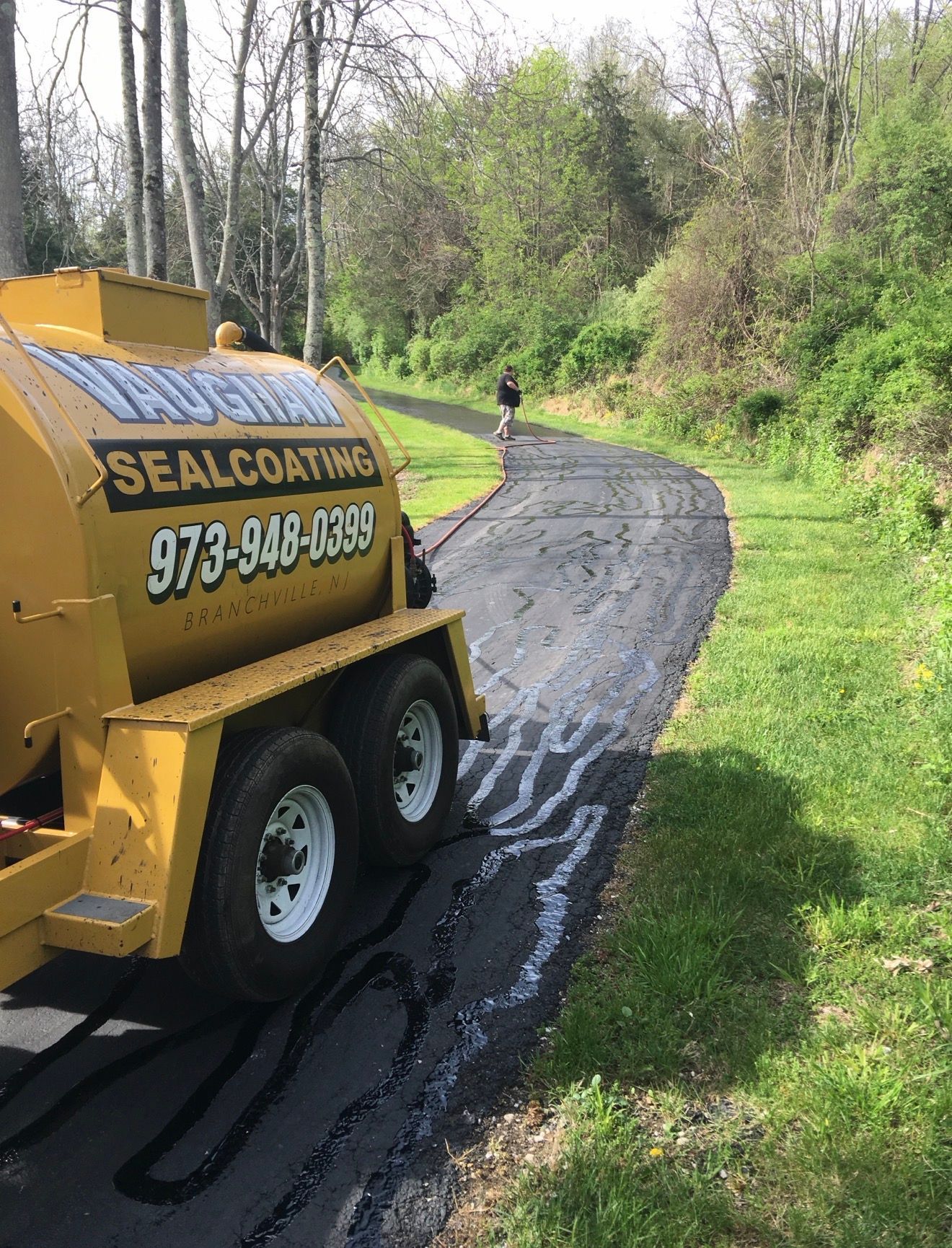 A yellow sealcoating trailer parked on a freshly paved, dark asphalt path bordered by green grass and trees.