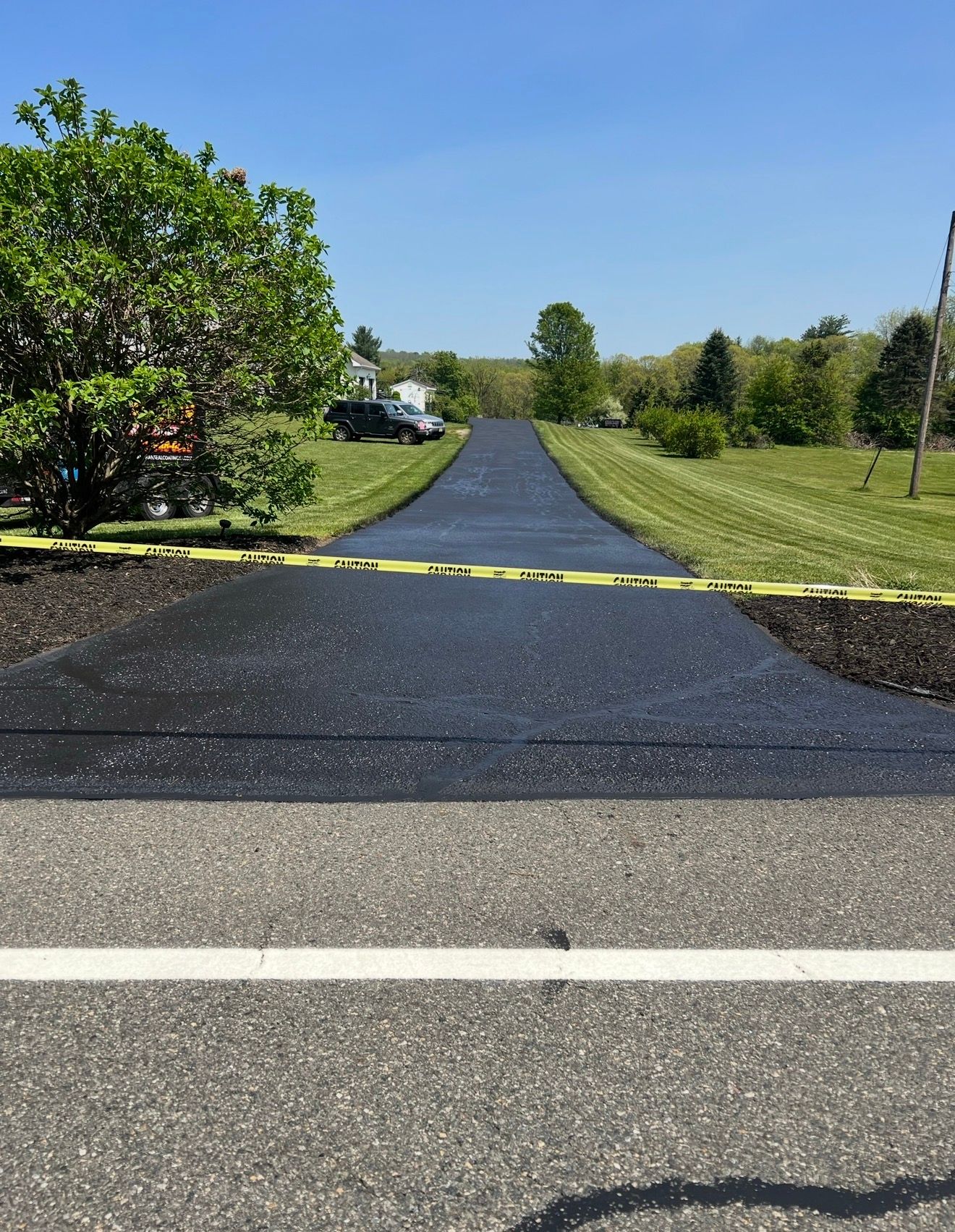 A freshly paved, black asphalt driveway leads from a paved road into a green, grassy yard under a clear blue sky.