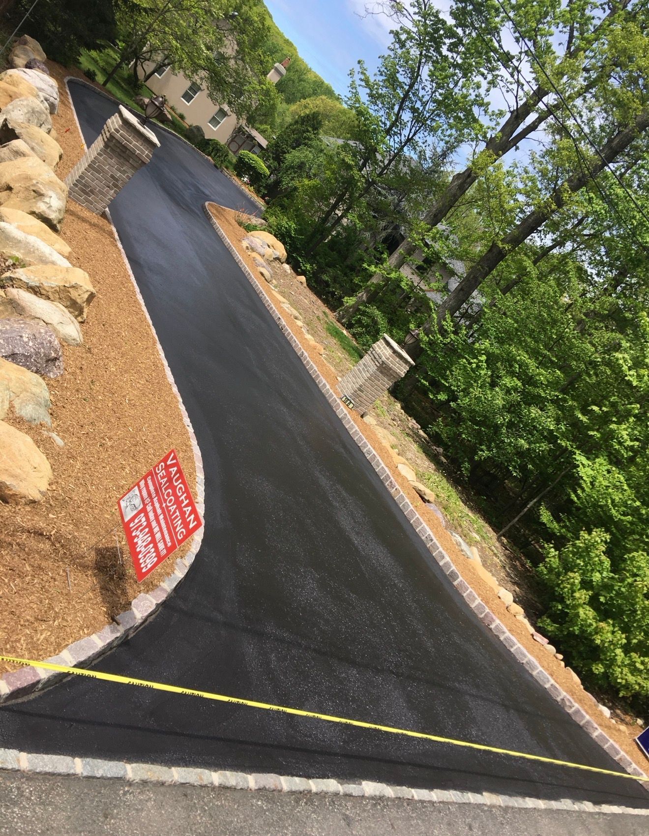 A freshly paved black asphalt driveway curves uphill, framed by stone pillars, stone borders, and a gravel embankment.