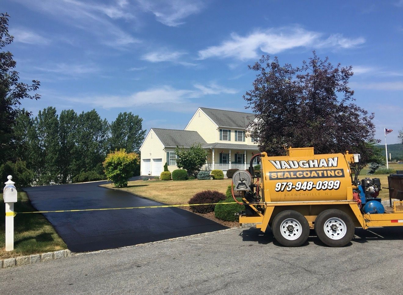 A yellow Vaughan Sealcoating trailer sits next to a freshly sealed black driveway leading to a house under a blue sky.