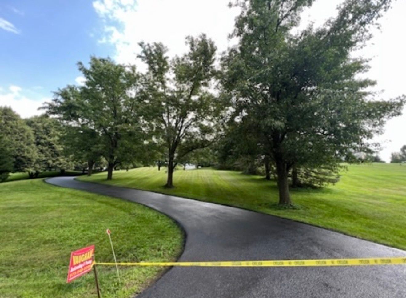 A paved, curving driveway surrounded by lush green lawns and large trees, blocked by a yellow caution tape.