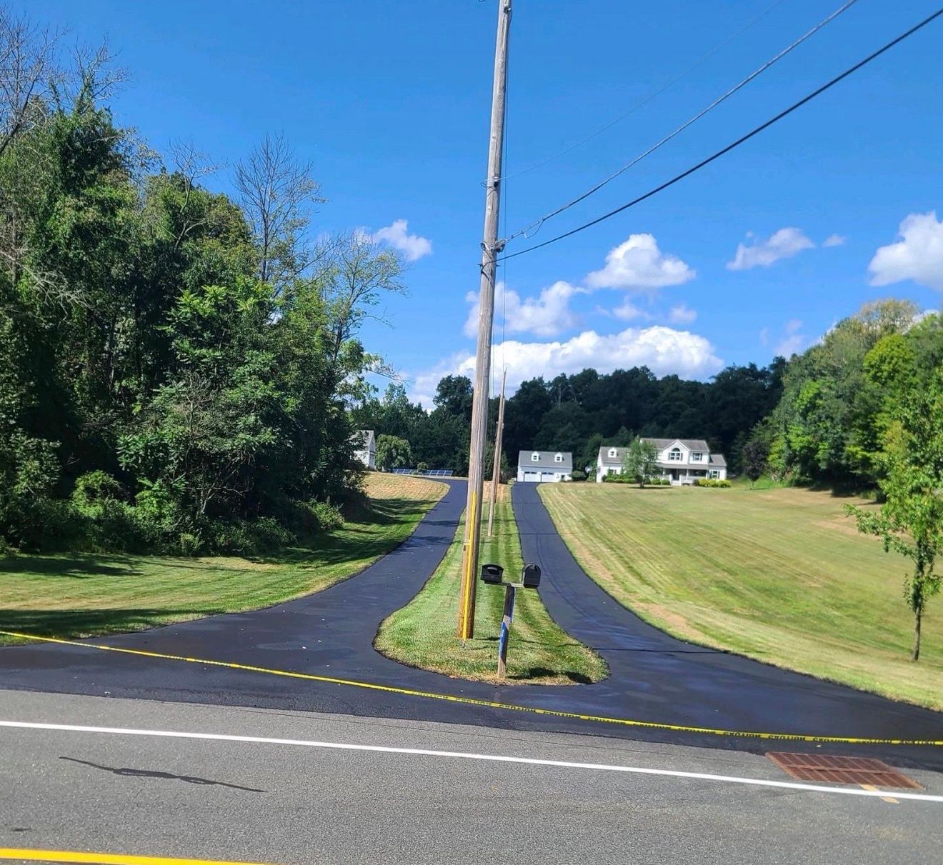 A split-entry paved driveway branches around a grassy median with a utility pole, leading toward houses under a blue sky.