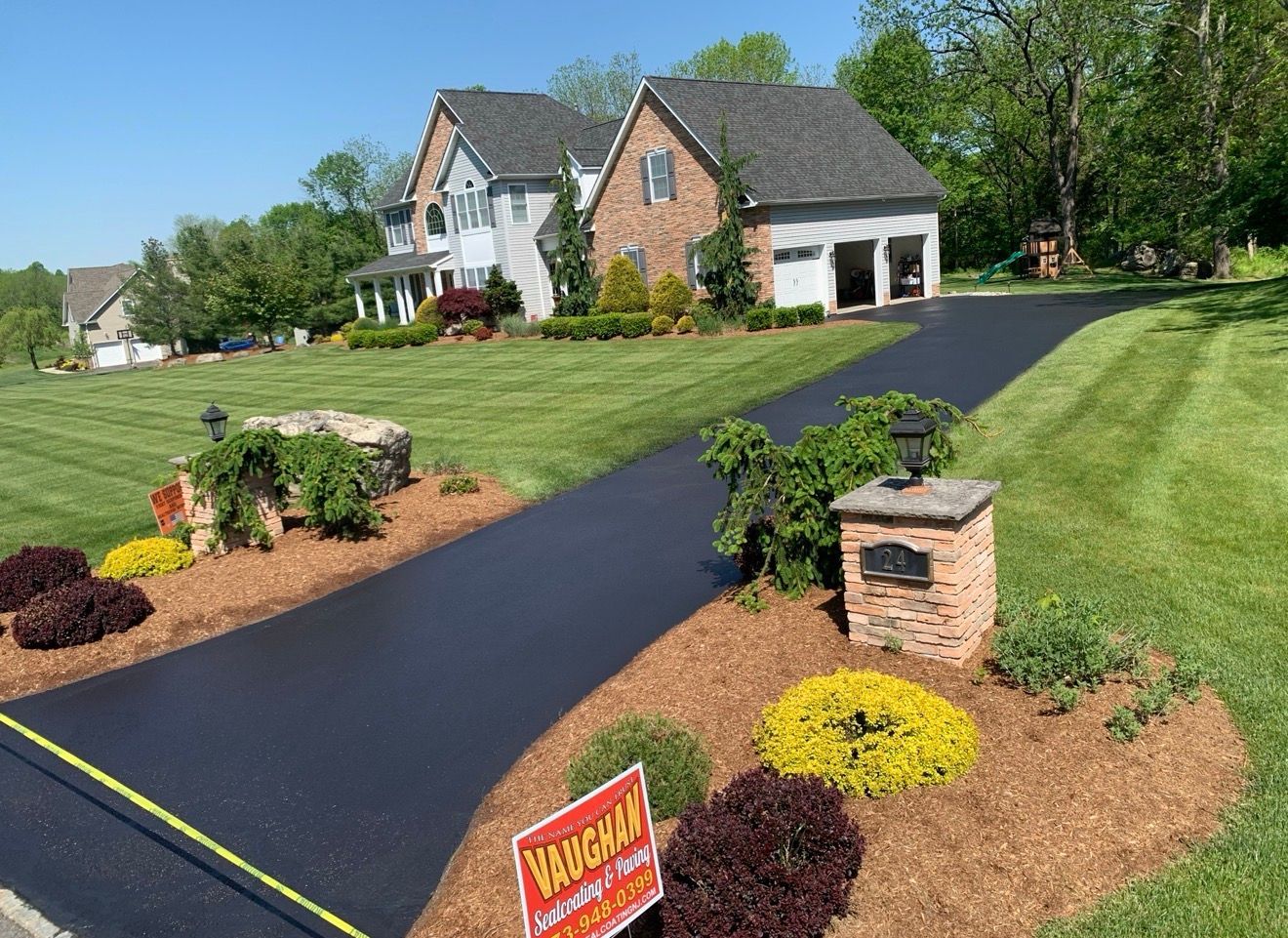 A paved driveway leads to a brick house on a grassy lawn with a landscaped garden bed containing flowers and a sign.