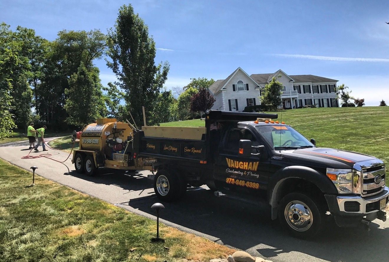 A black dump truck towing a yellow trailer on a residential driveway near a large white house on a sunny day.