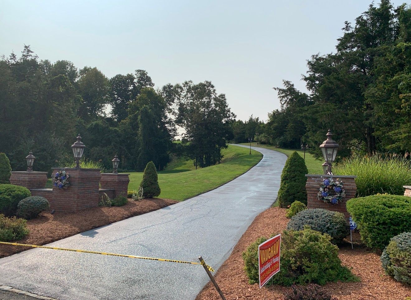 A winding gravel driveway flanked by brick pillars and green landscaping, blocked by caution tape on a sunny day.