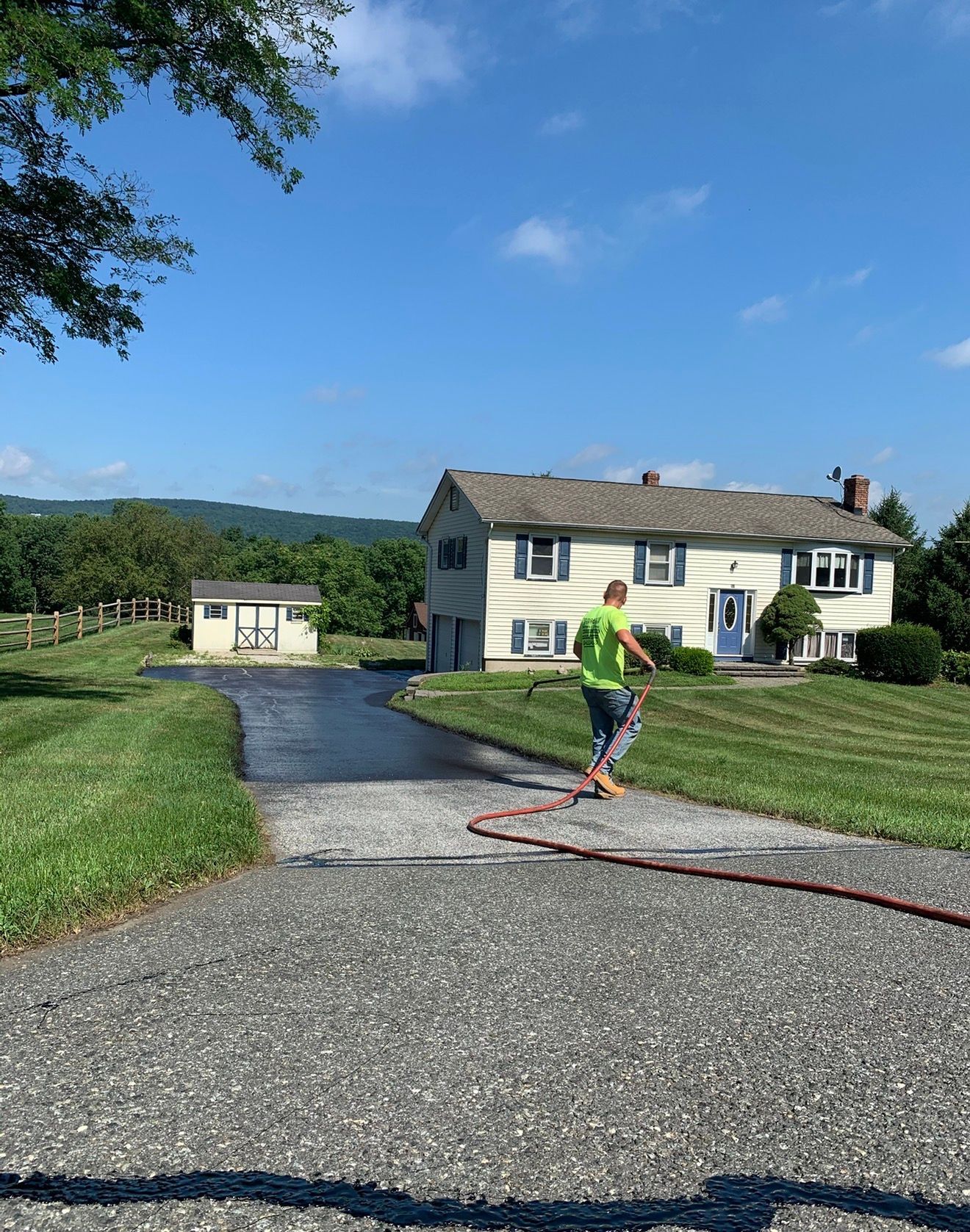 A worker in a neon green shirt sprays sealant on a gravel driveway in front of a house on a sunny day.