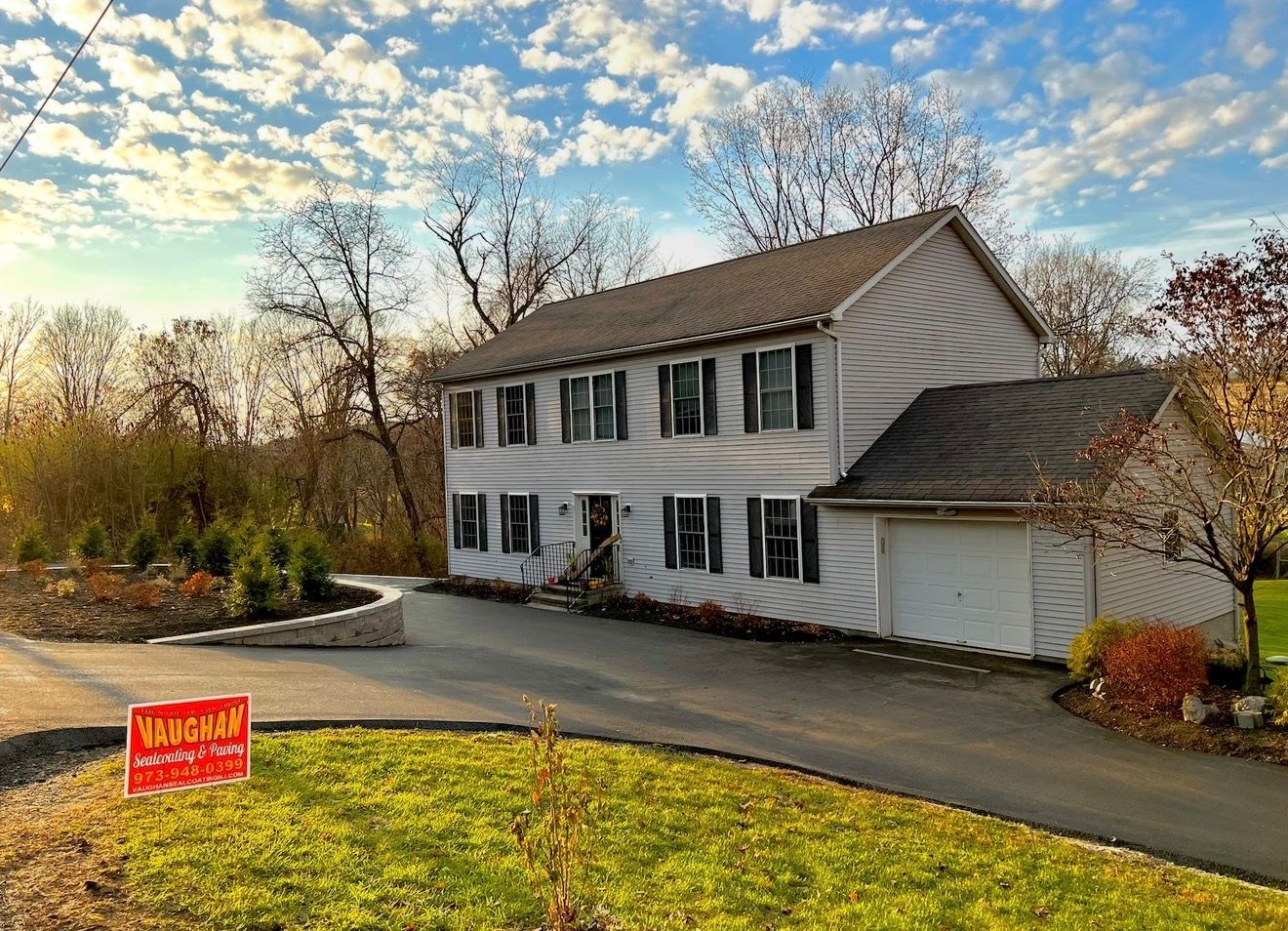 A two-story white house with dark shutters and a garage stands next to a curved driveway with a red real estate sign.