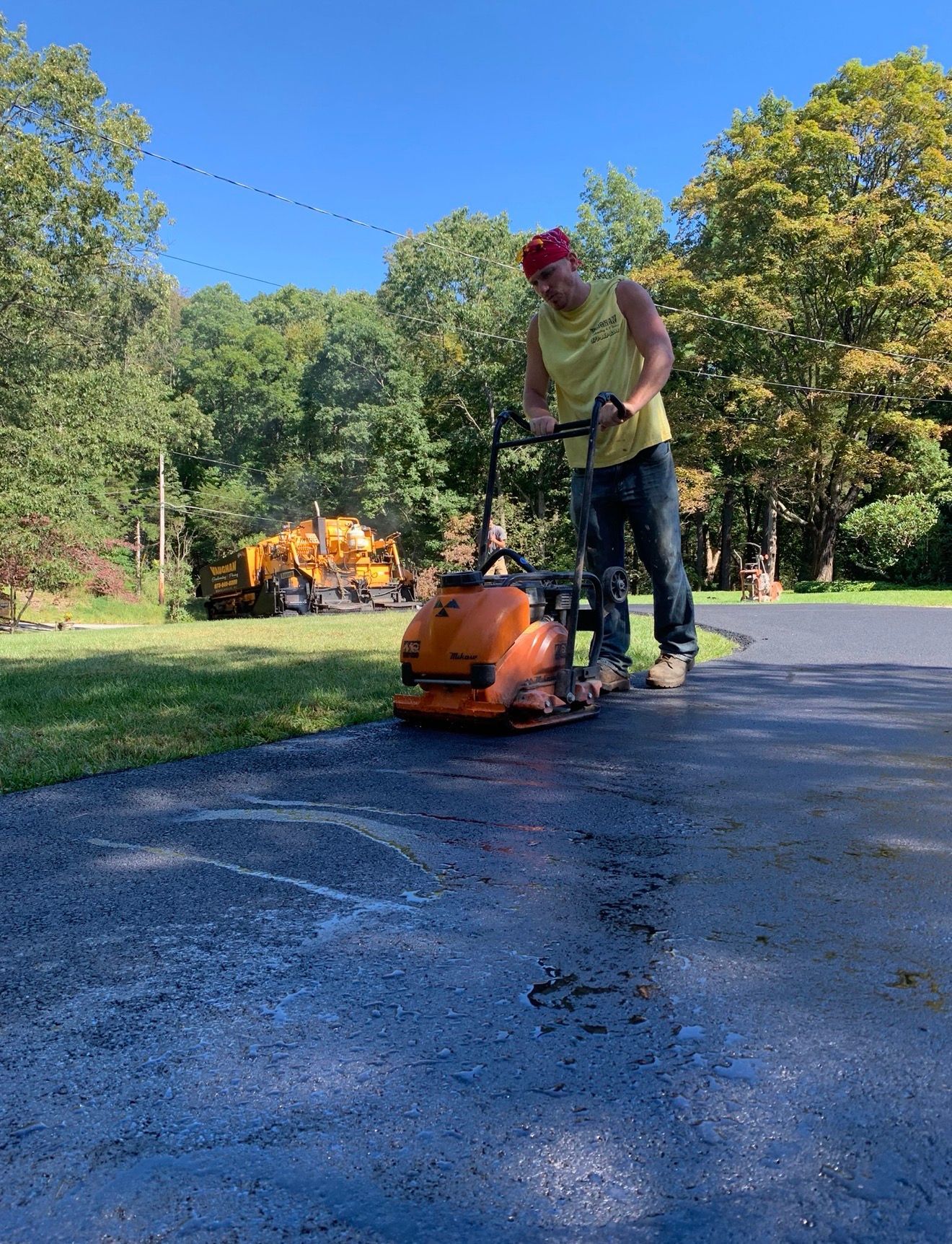 A person using an orange plate compactor to smooth freshly laid asphalt on a sunny driveway, with trees in the background.