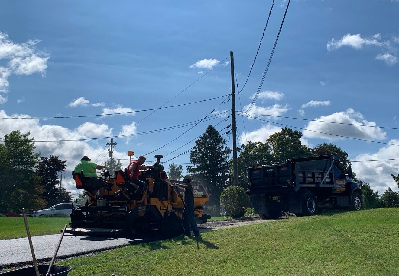 Construction crew members operate an asphalt paving machine on a gravel driveway, with a dump truck nearby under a sunny sky.
