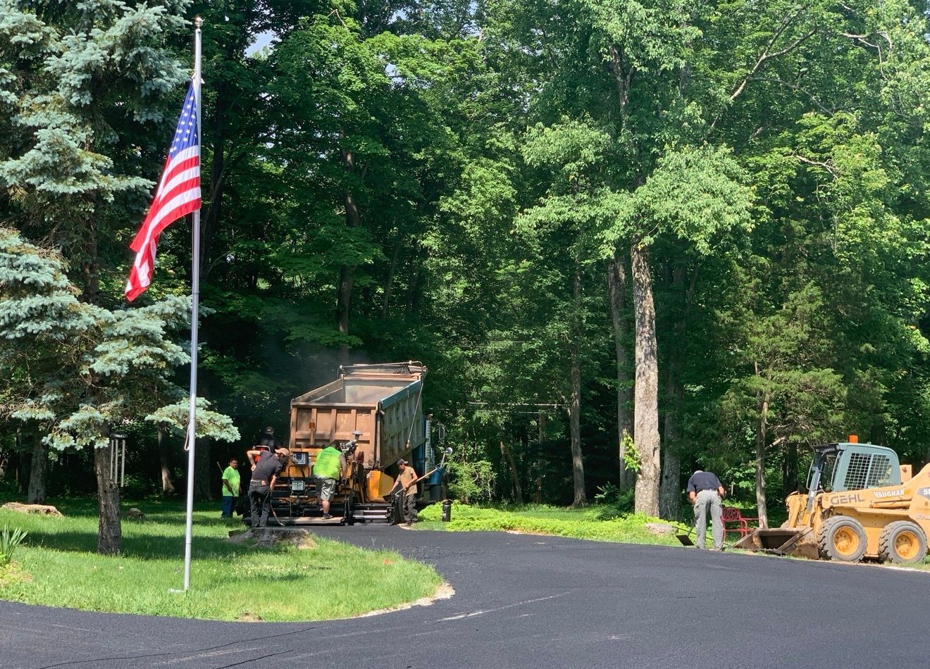 Workers operate heavy machinery to pave a driveway with asphalt near a forest, with an American flag flying nearby.