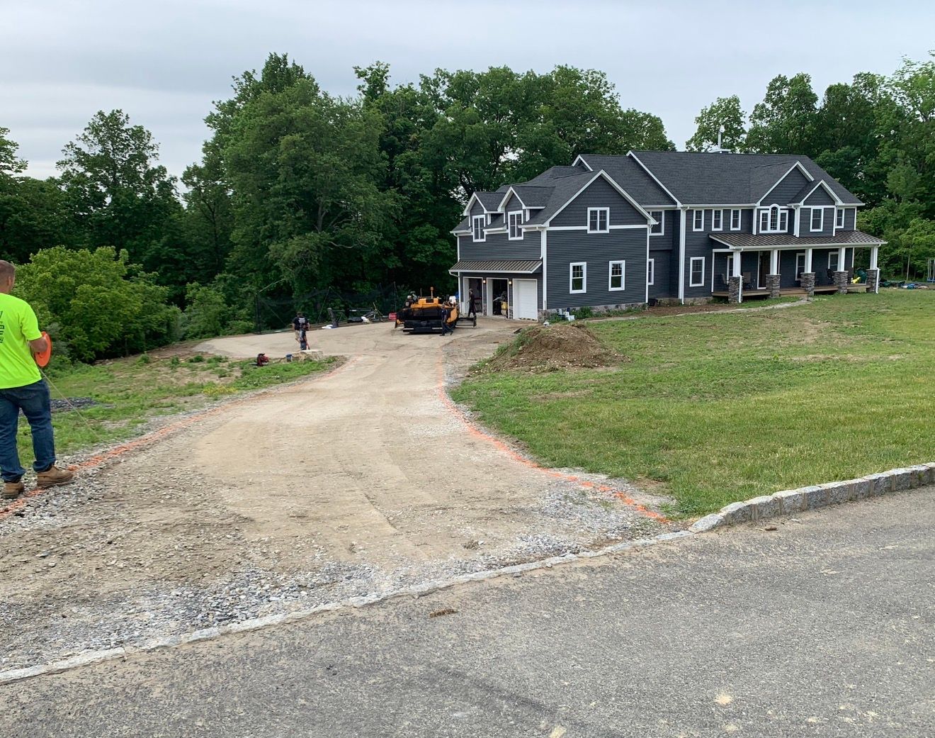 A gravel driveway leads to a large, dark-sided two-story house under construction in a grassy field bordered by trees.