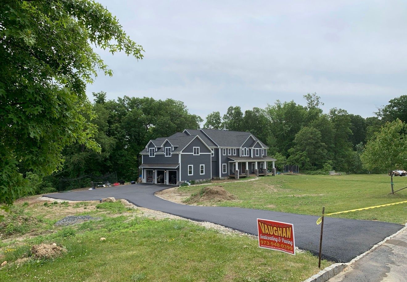 A dark grey two-story house sits at the end of a long, newly paved black driveway, surrounded by green trees and grass.