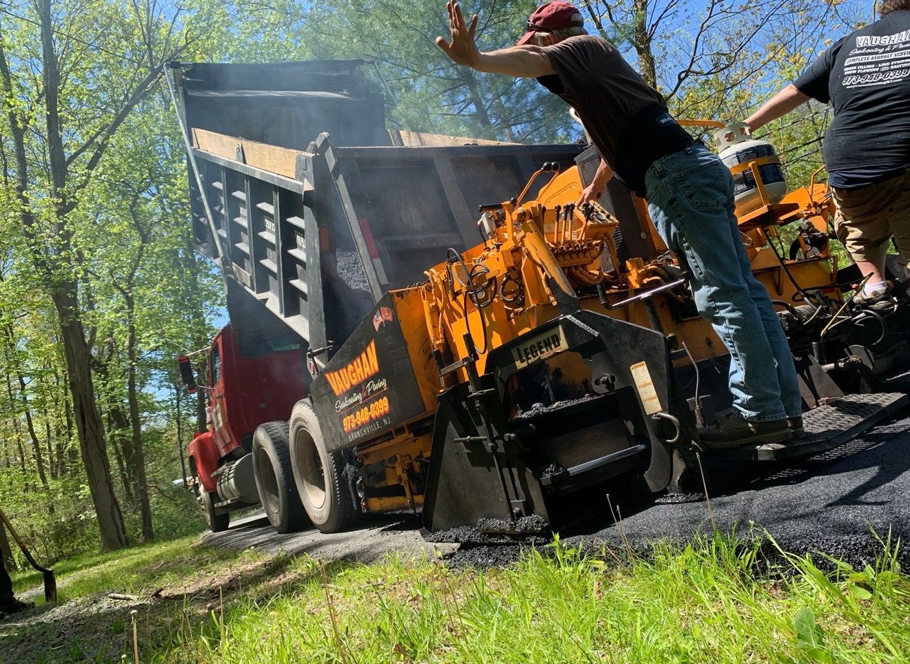A construction worker guides a red dump truck as it empties material into a yellow asphalt paving machine in a wooded area.