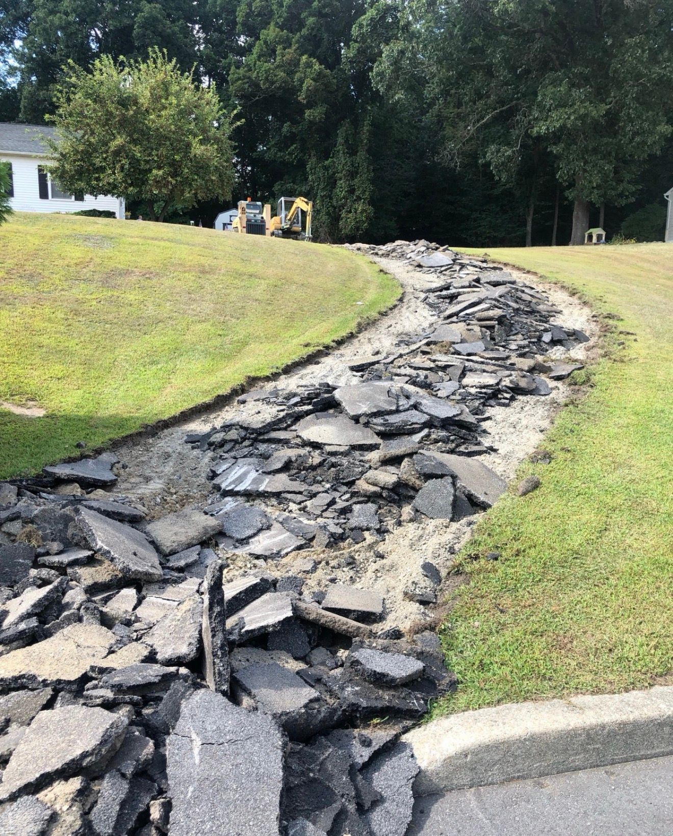 A damaged asphalt driveway being torn up, with loose chunks of debris lining the path in a residential yard.