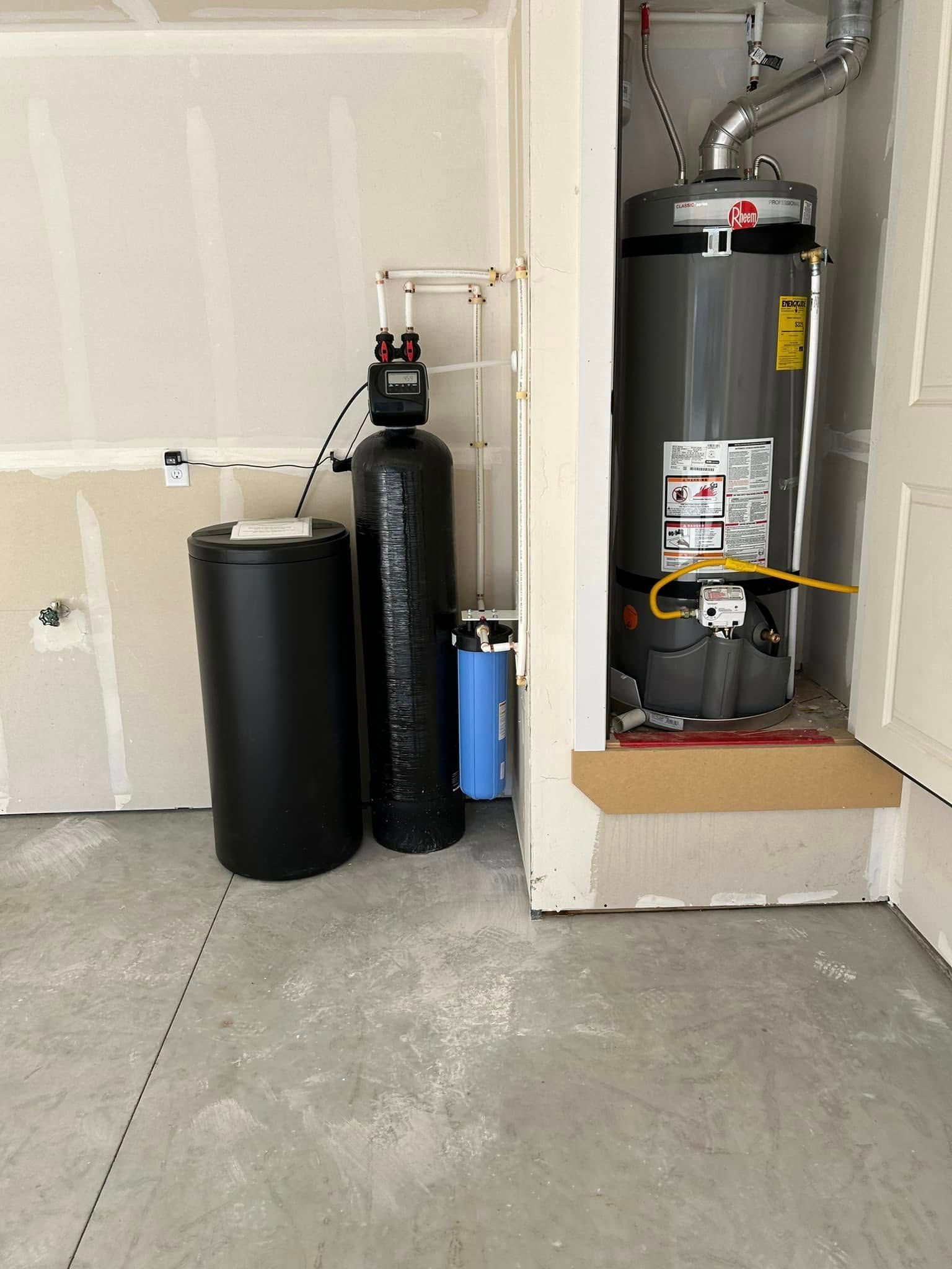 Water softener system, filter, and water heater in a garage setting. Black tanks and grey water heater.