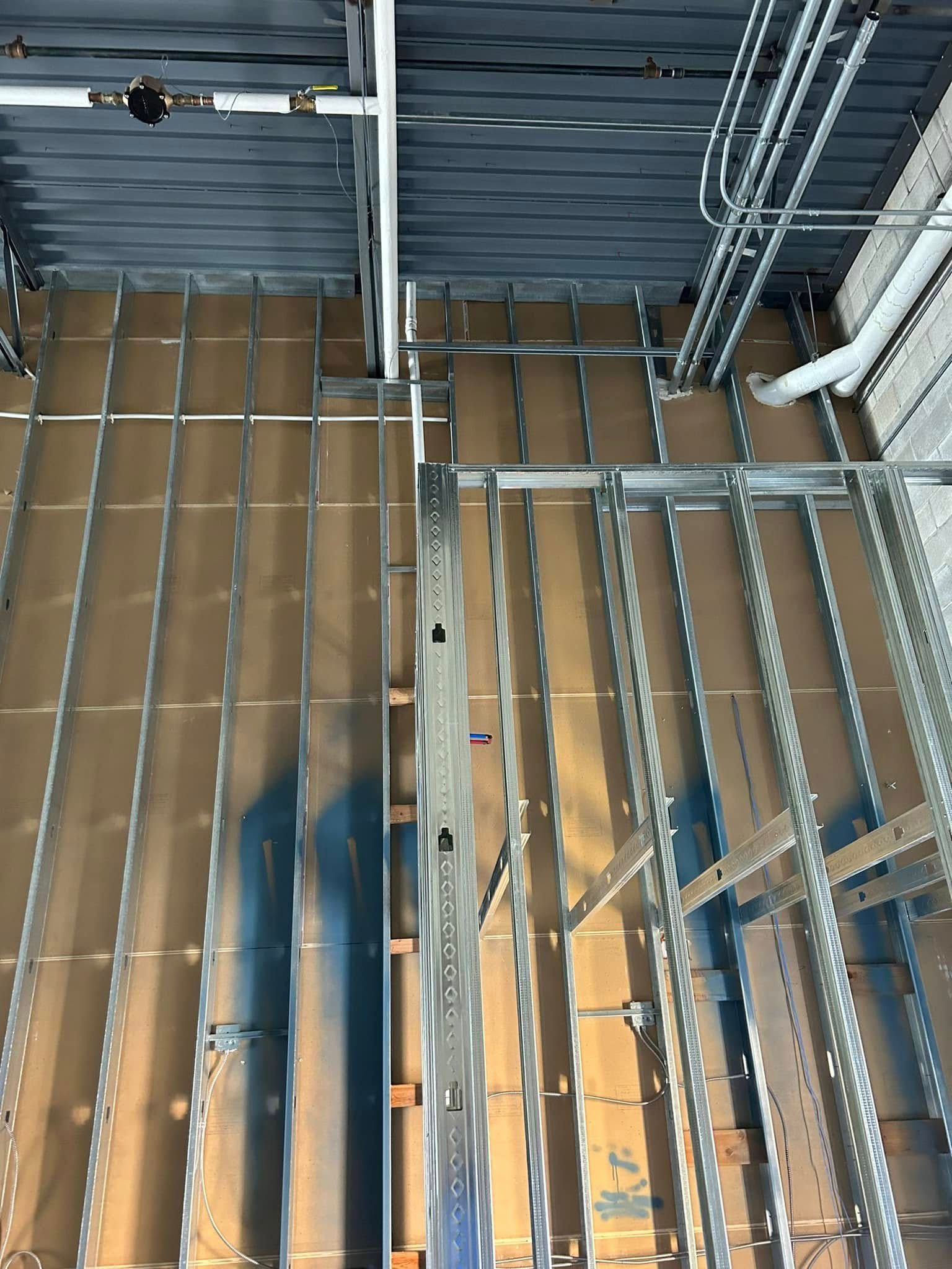 Construction site interior: metal framing, brown insulation, exposed pipes, and conduits in the ceiling and walls.