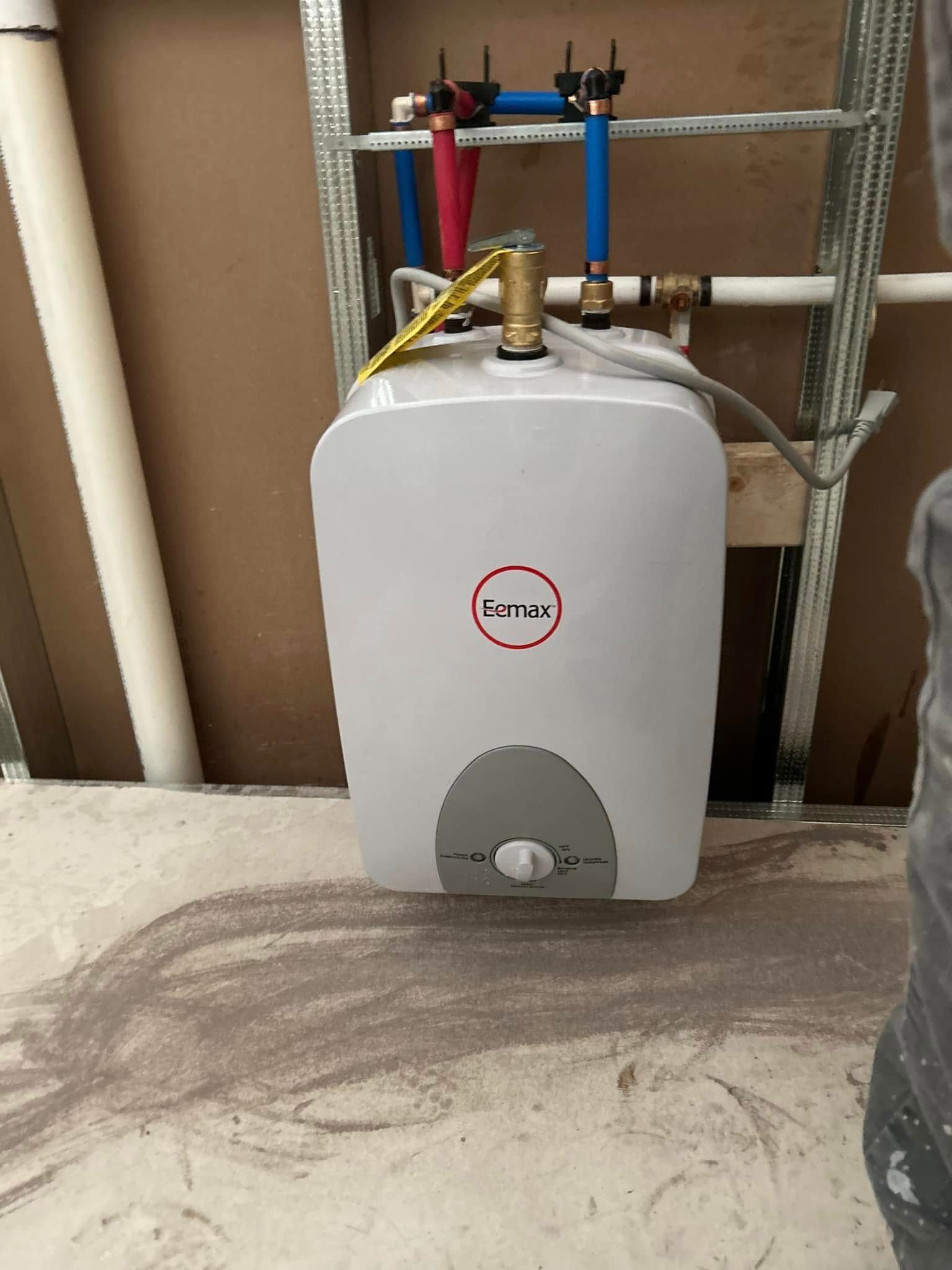 A small, white, electric water heater installed in a wall with plumbing and electrical connections visible.