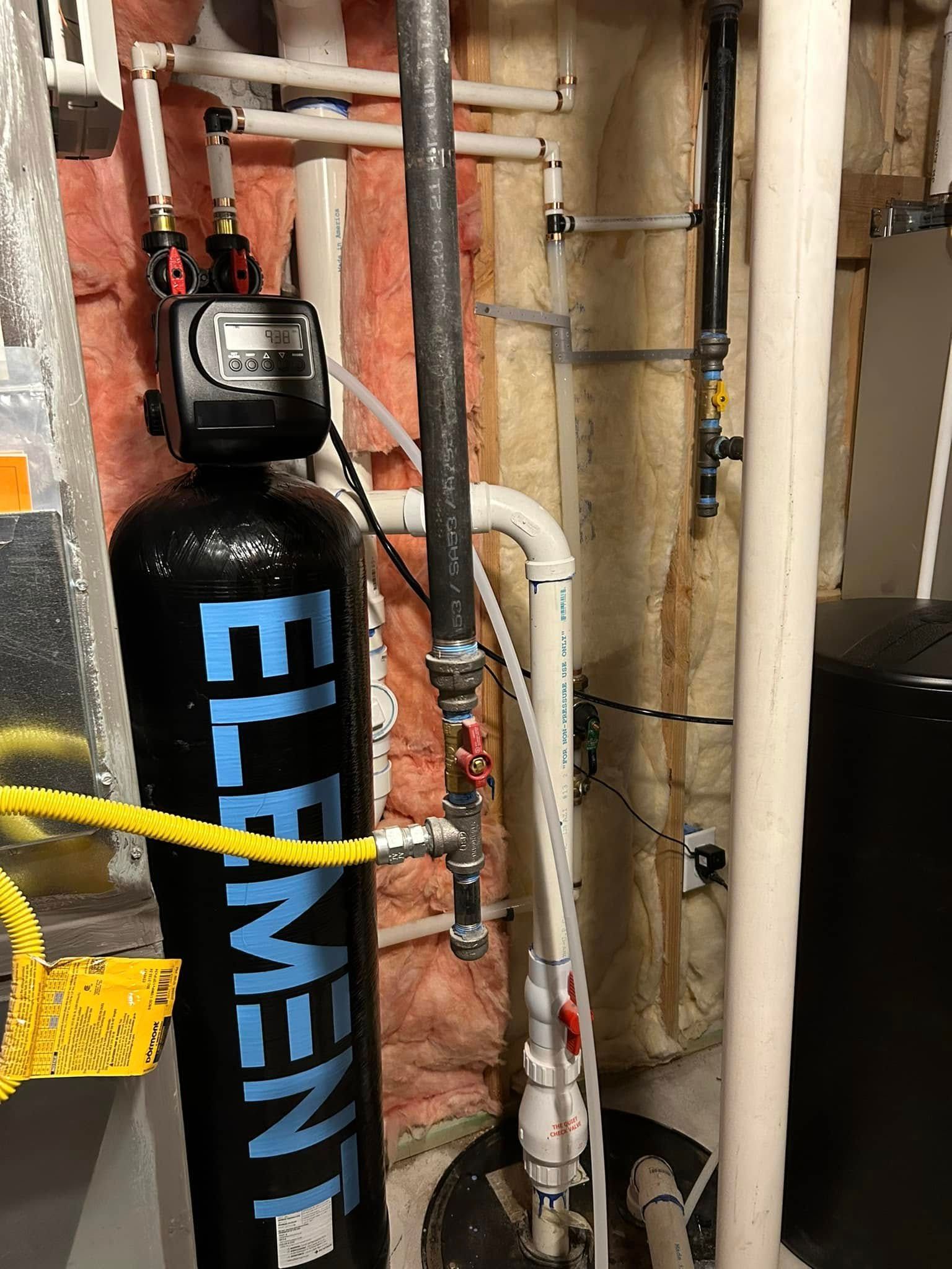 Water filtration system in a basement. A tall black tank has an electronic display and is connected to white and black pipes.