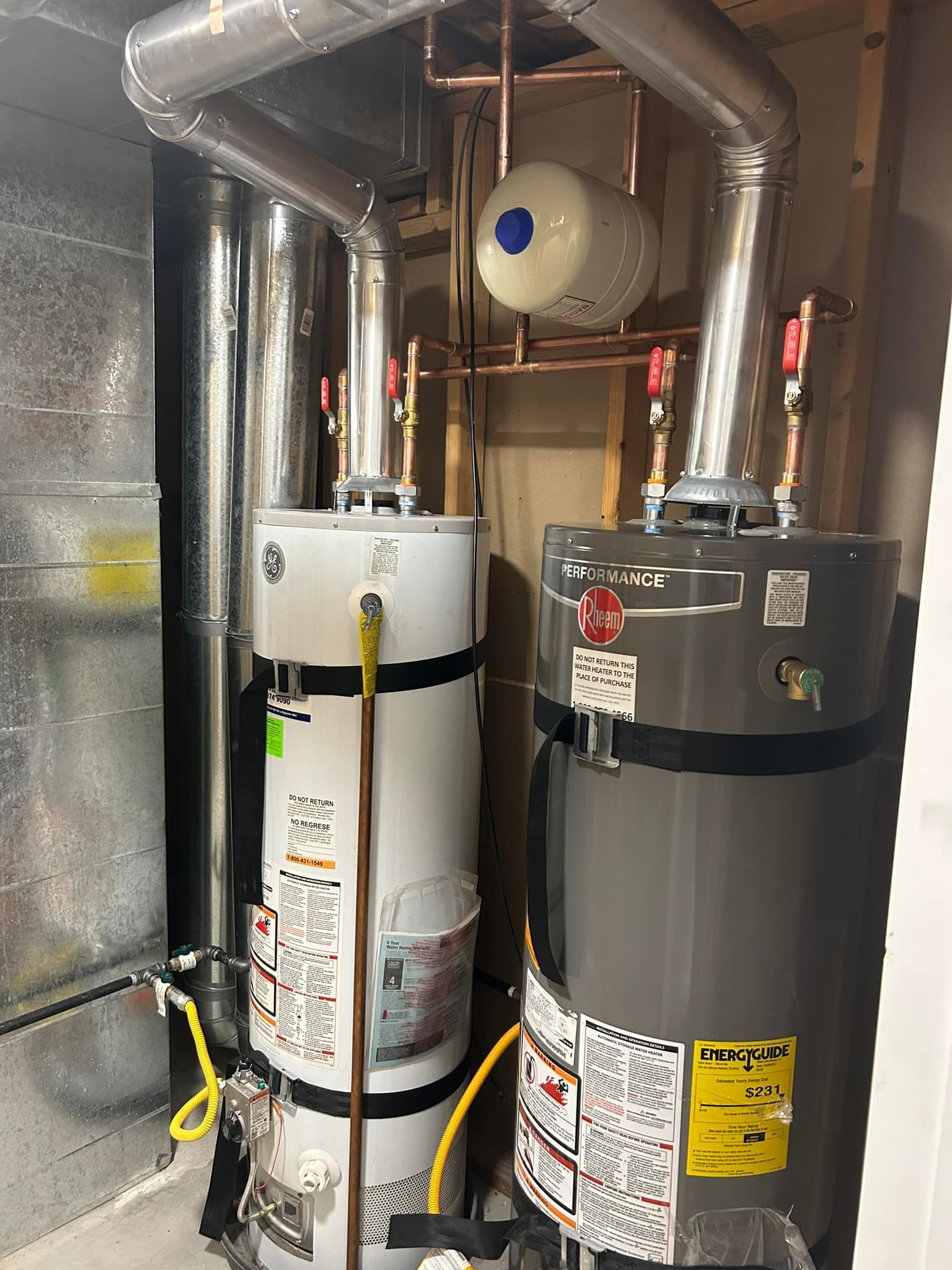 Two water heaters with copper pipes and a white expansion tank in a utility room.