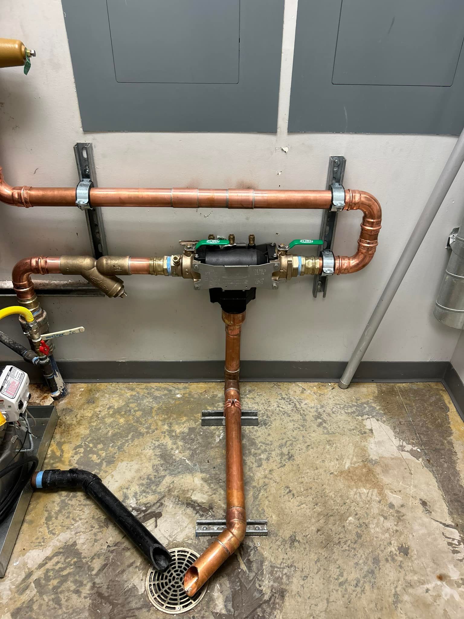 Copper pipes and fittings, mounted on a wall. Includes a valve, and a floor drain at the bottom.