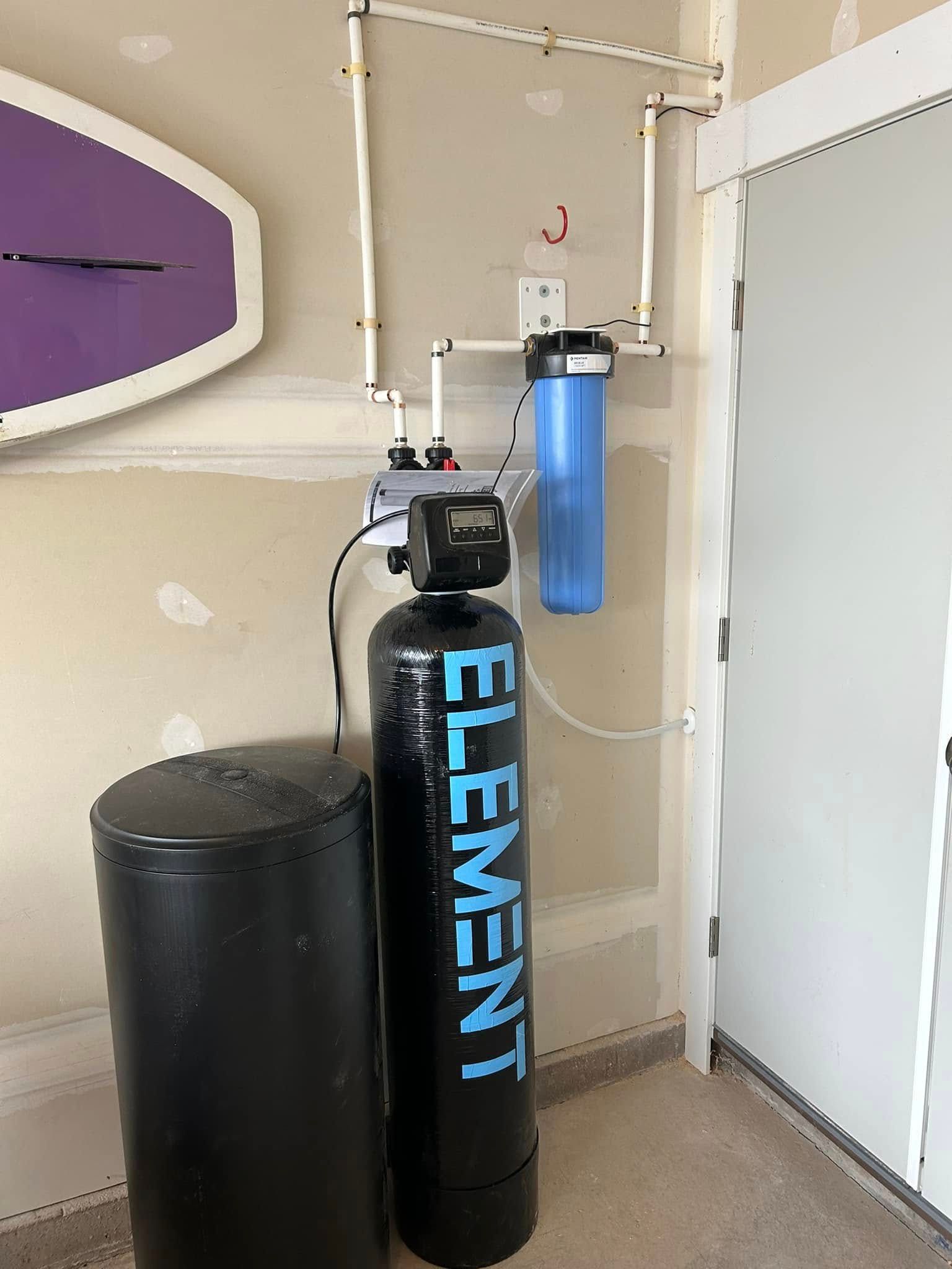 Water filtration system in a garage: black tank and filter, blue filter, white pipes.