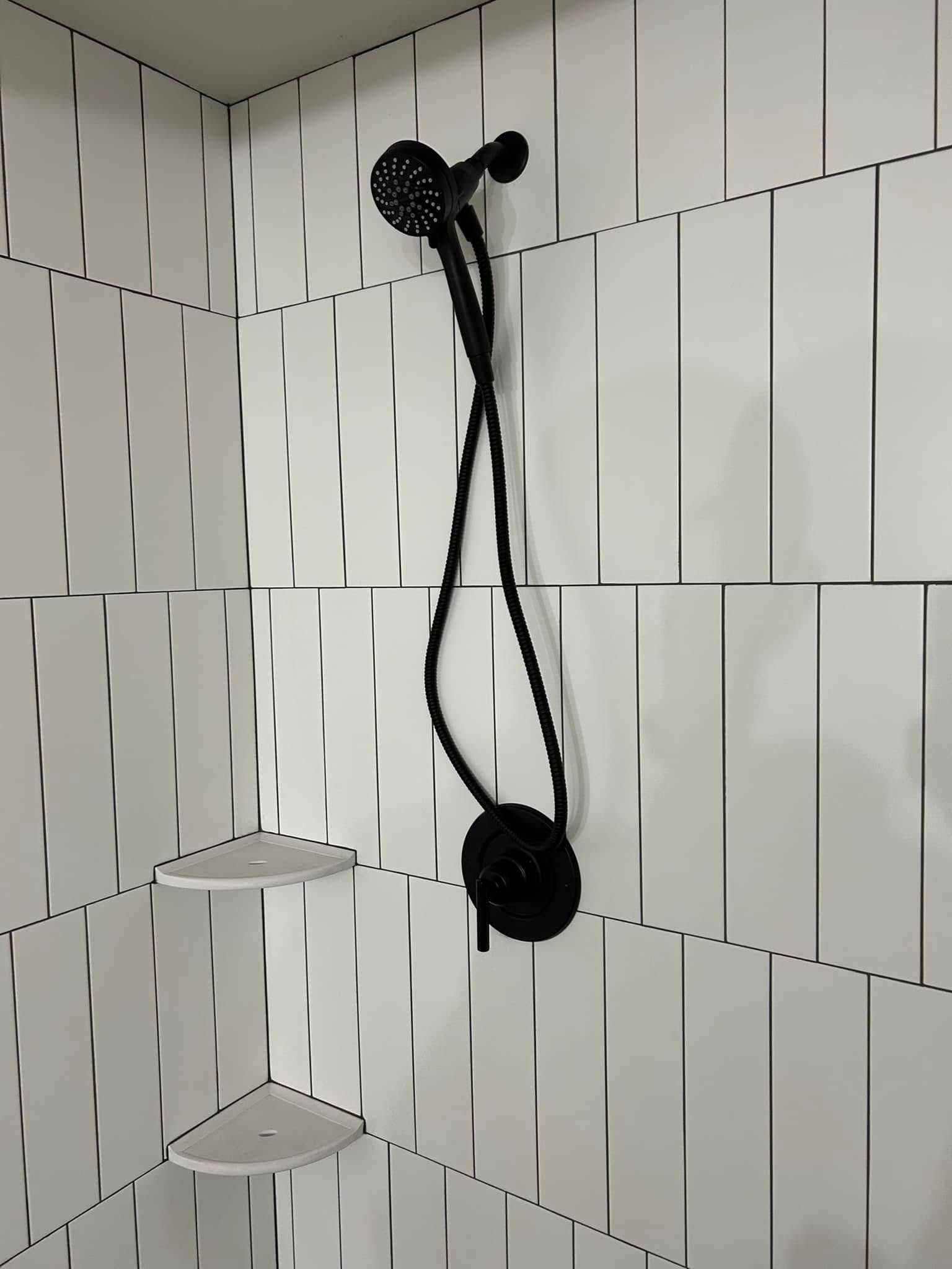 Black shower head and controls on white tiled wall with two corner shelves.