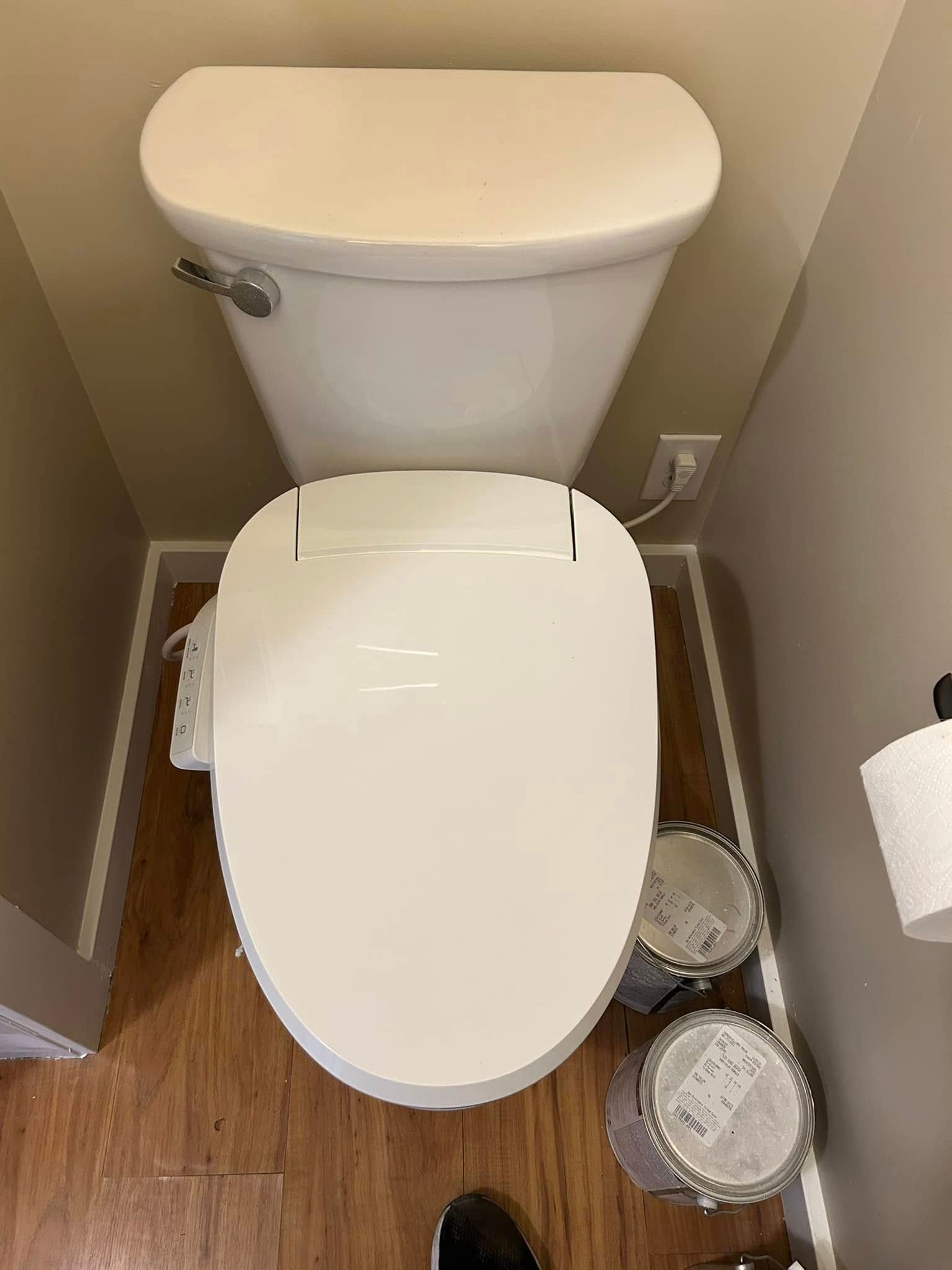 White toilet in a small bathroom with a wood floor, two paint buckets, and a roll of toilet paper.
