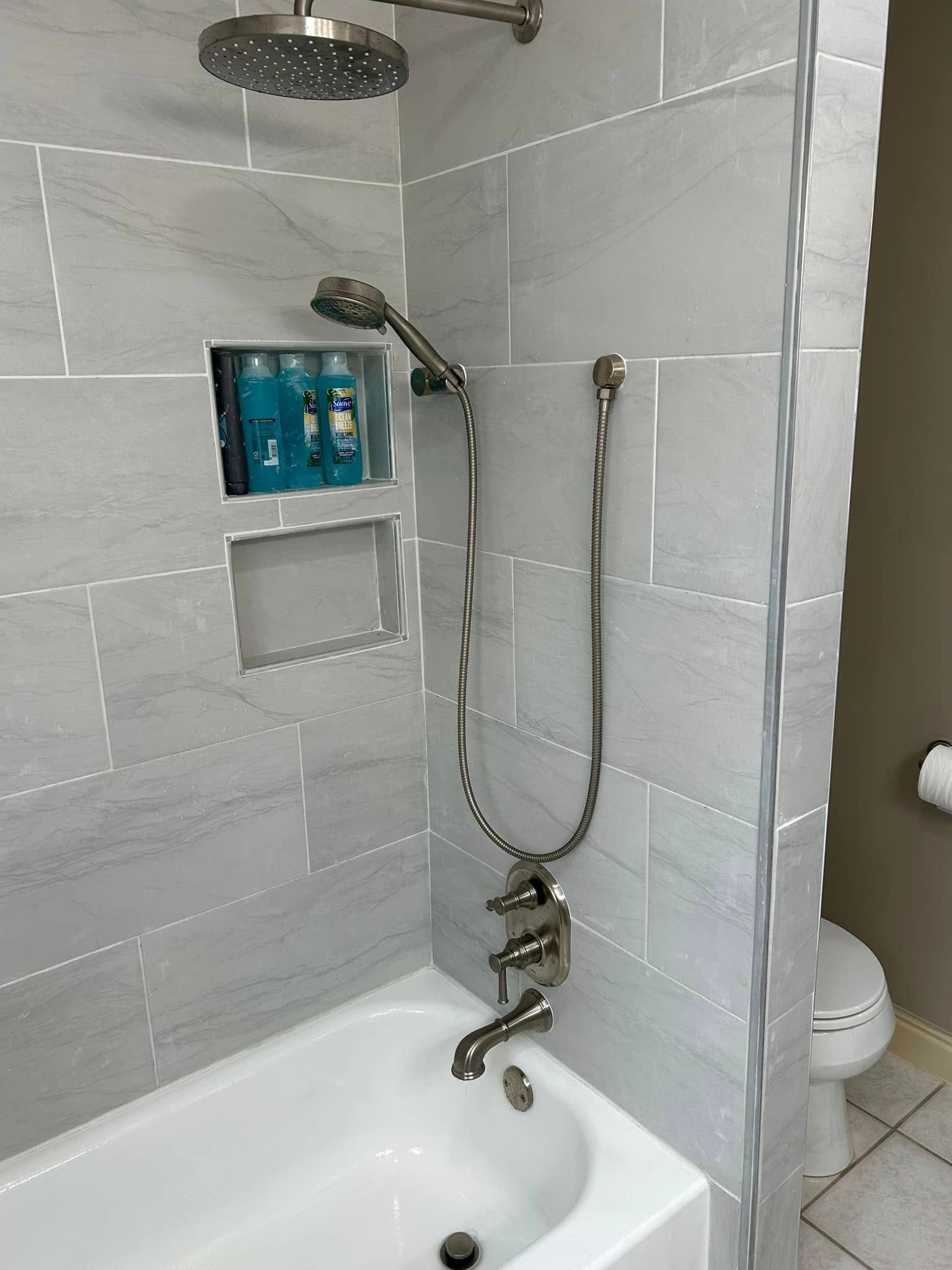 Bathroom with white tub, light gray tiled walls, showerhead, and toiletries in a recessed shelf.