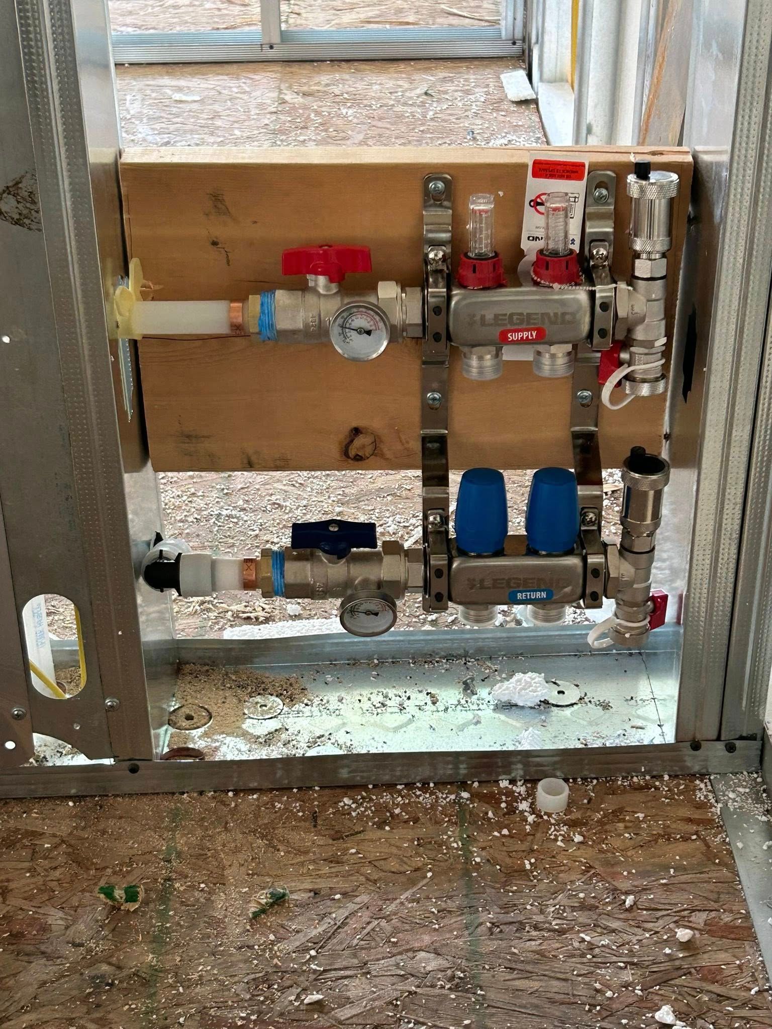 Plumbing manifold with red, blue, and silver pipes, valves, and gauges mounted on wood.