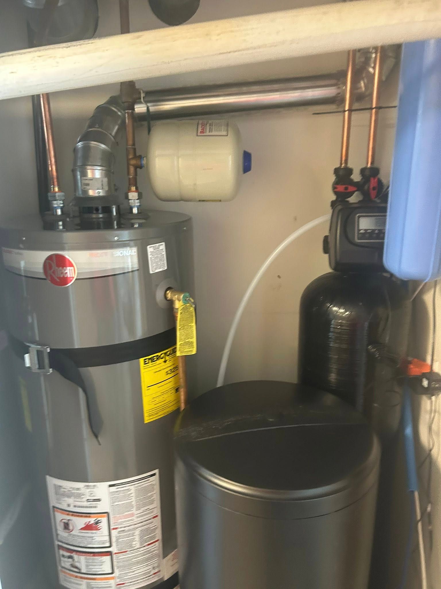 Water heater, expansion tank, and water softener in a utility room. Copper pipes and black and gray tanks.