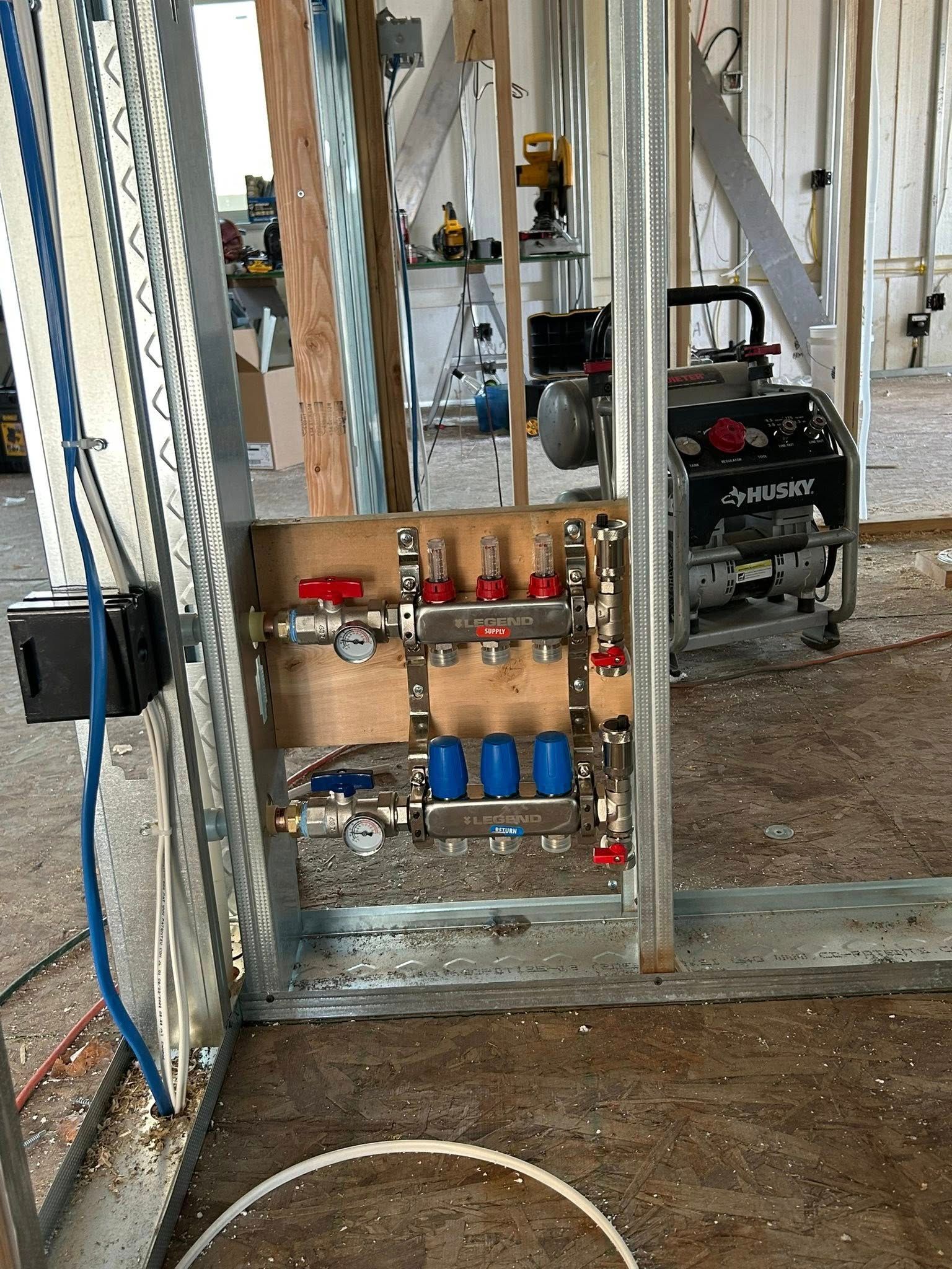 Framed construction with radiant heat manifold; red & blue valves, pipes, and generator in background.