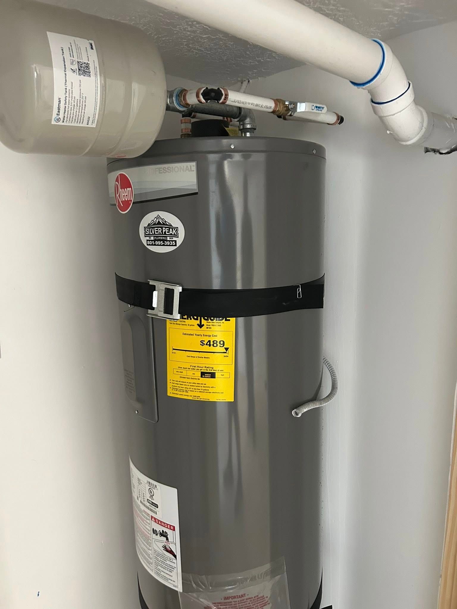 A tall, gray water heater with black strap, white pipes, and a small water tank in a white room.